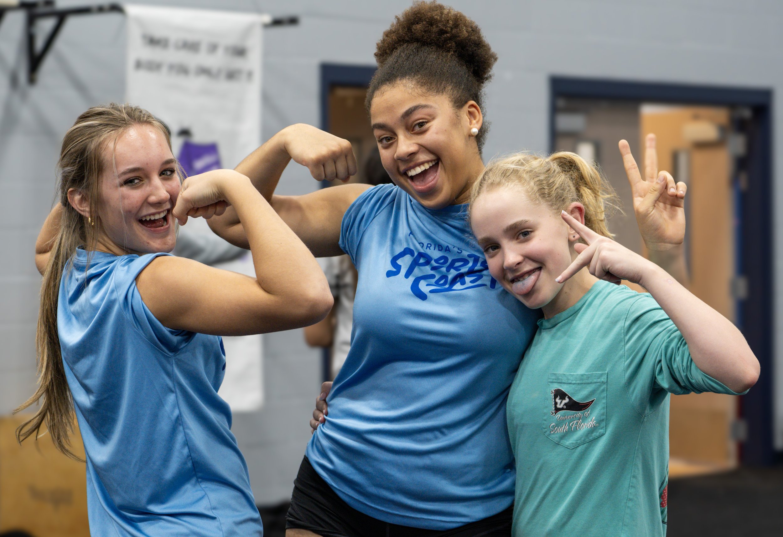 Three young women in matching teal and blue sports T-shirts in a gym, posing playfully and smiling for the camera.