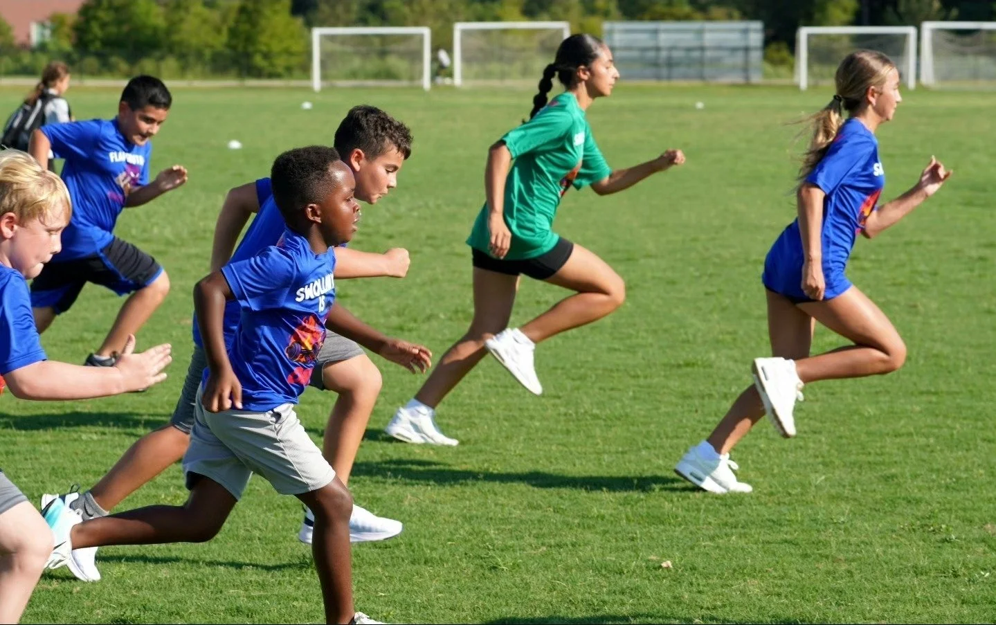 Children running on a grassy field during daytime, some wearing blue shirts and one girl wearing a green shirt.