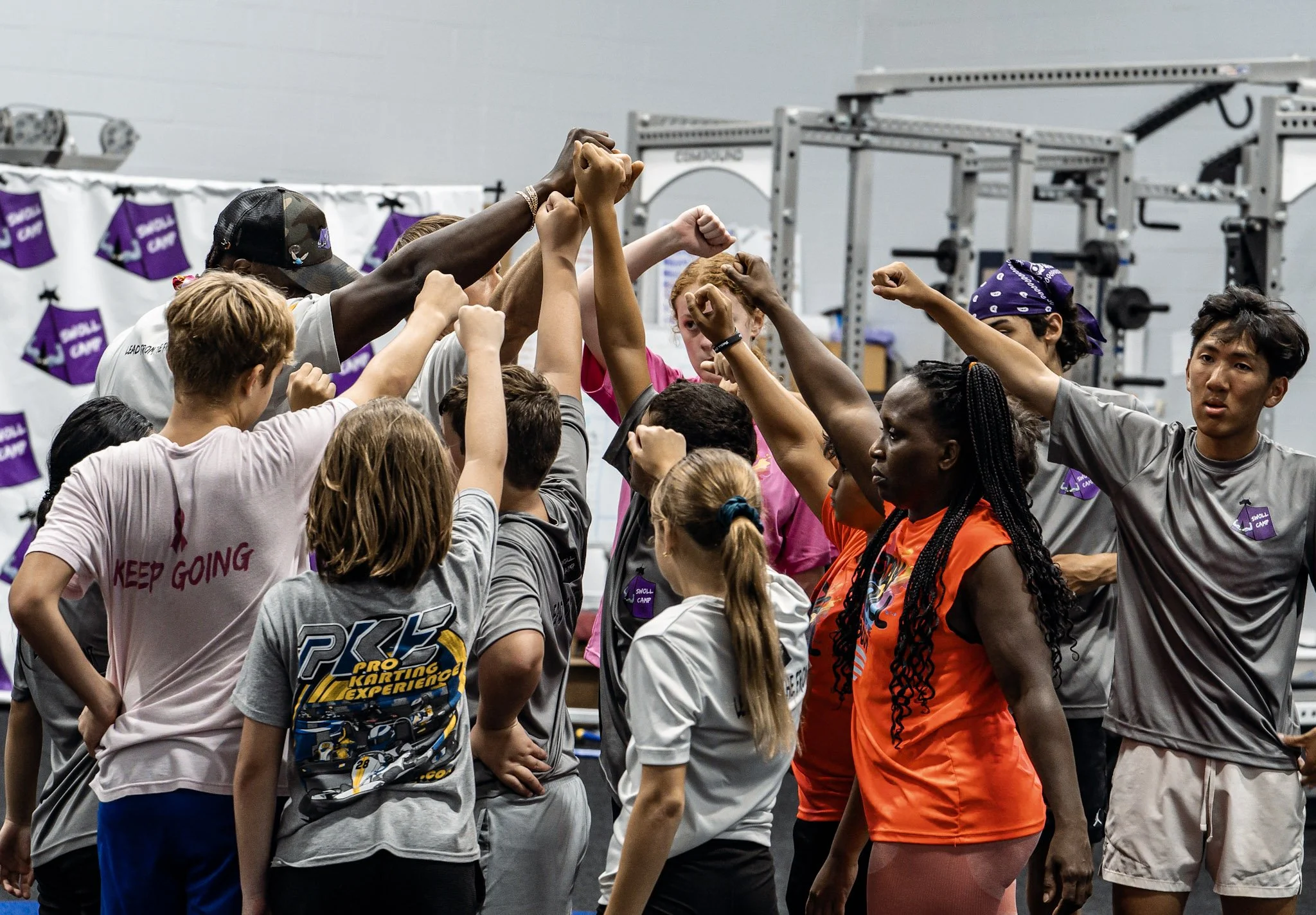 A diverse group of people forming a huddle with fists raised inside a gym or sports facility, with weightlifting equipment in the background.