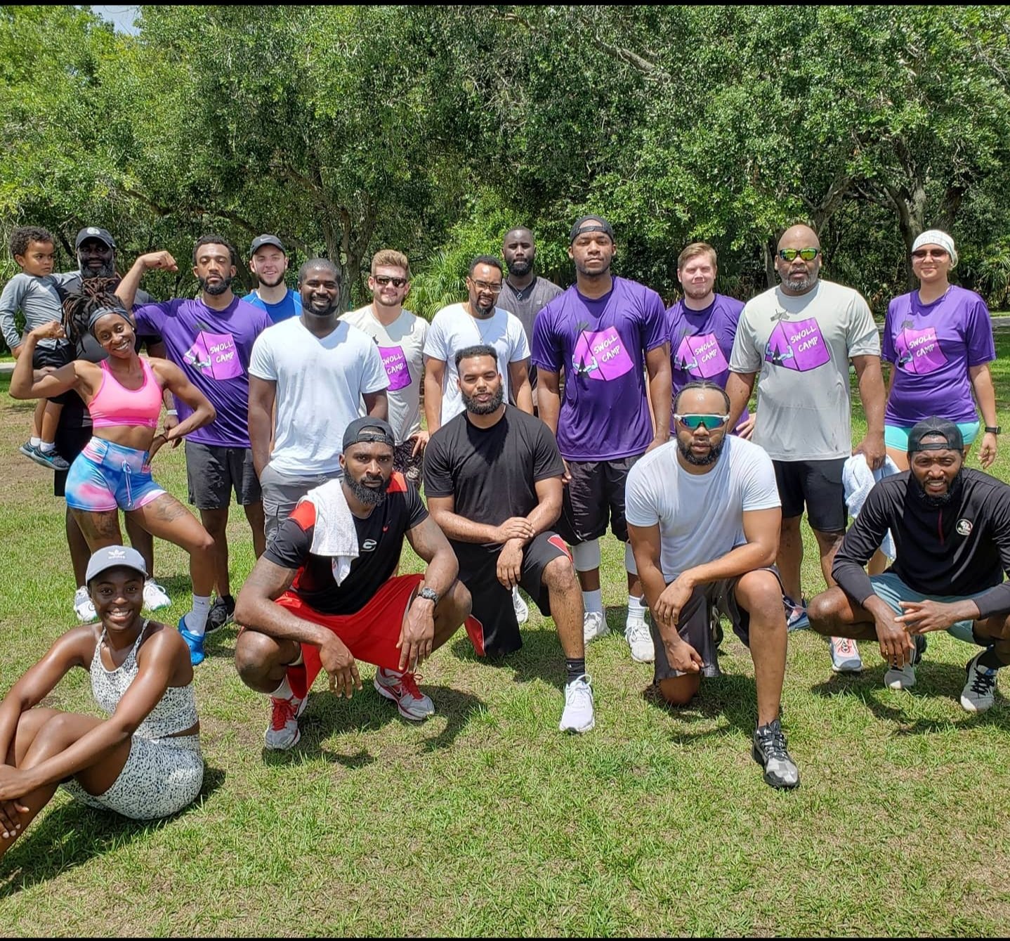 Group of people outdoors in a park, some wearing athletic clothing, posing for a photo on a sunny day.