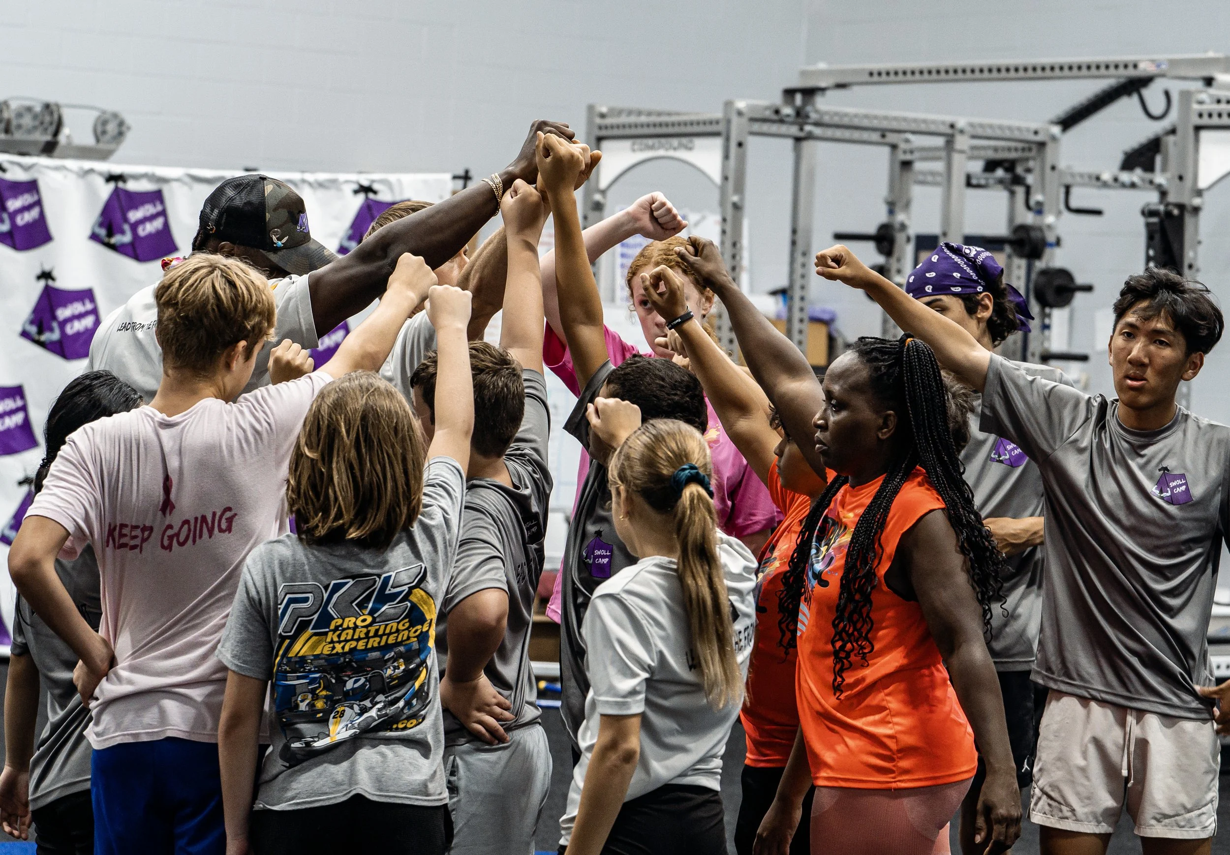 Group of diverse young people in a gym, forming a circle with their arms raised and hands stacked together in a sign of unity and teamwork.