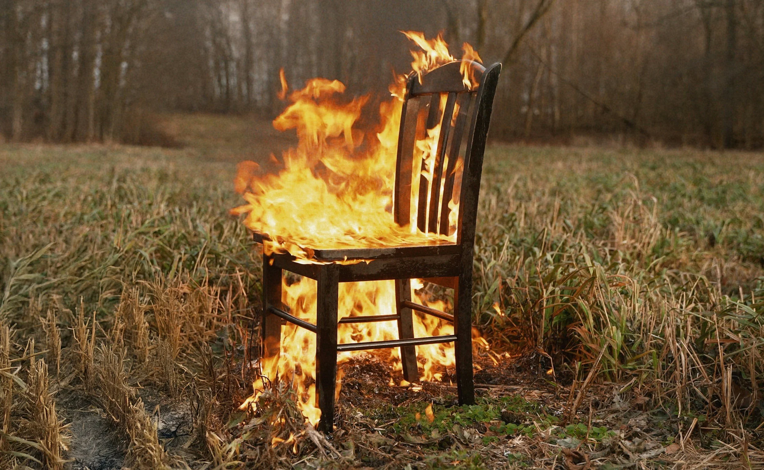 A wooden chair engulfed in flames in an open field with tall grass and wooded area in the background.