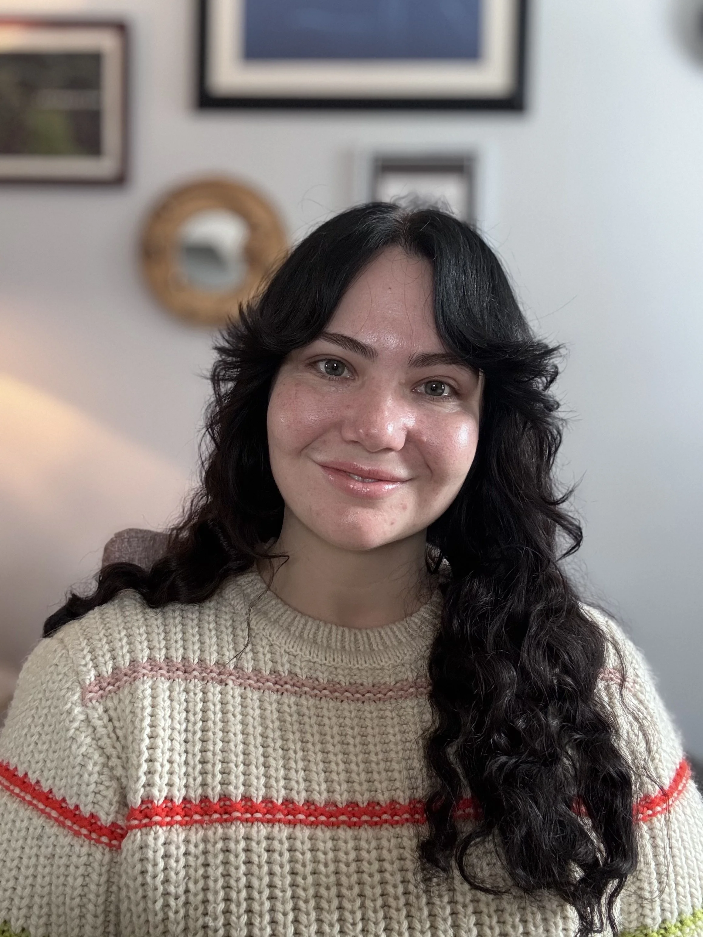 Close-up of a woman with long, dark, wavy hair, smiling, wearing a beige knit sweater with red and pink patterns, in front of a wall decorated with framed pictures and a circular mirror.