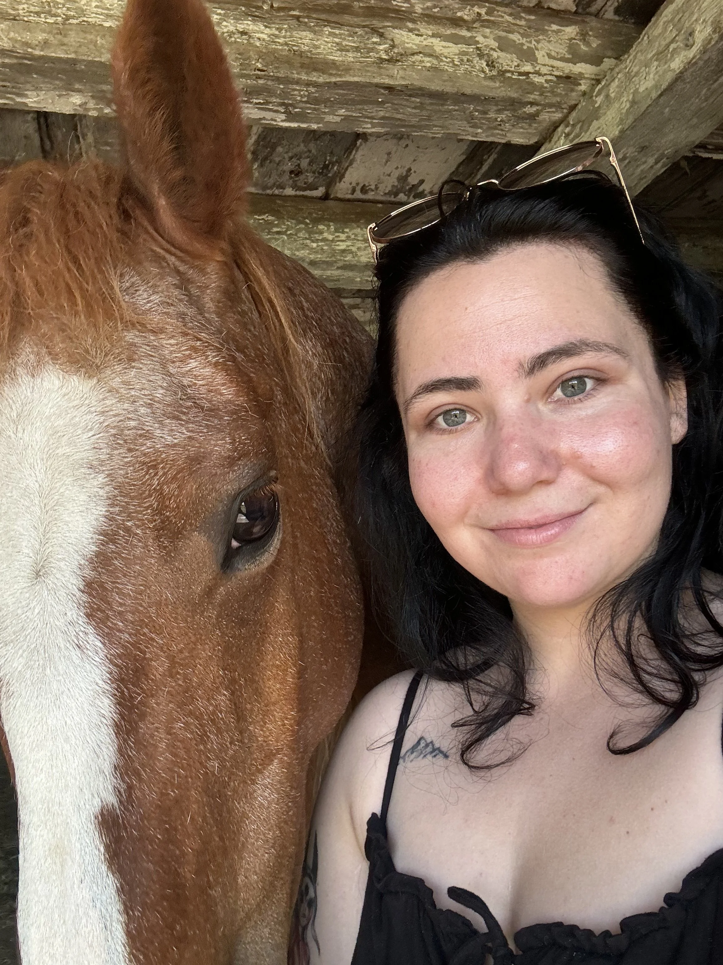 A woman with black hair and sunglasses on her head taking a selfie with a horse in a barn.