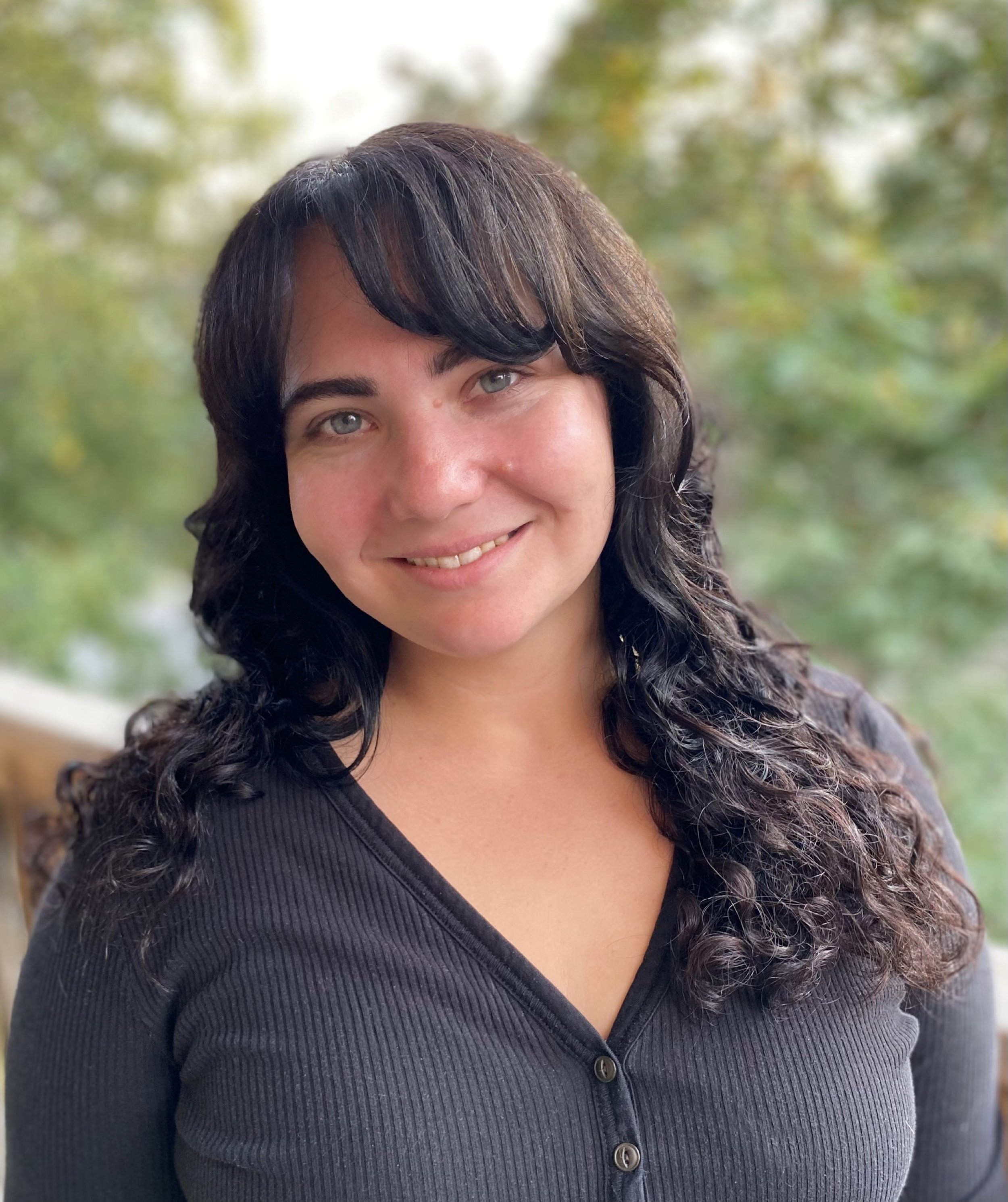 Close-up of a woman with black, curly hair and blue eyes, smiling outdoors with green trees in the background, wearing a black ribbed button-up shirt.