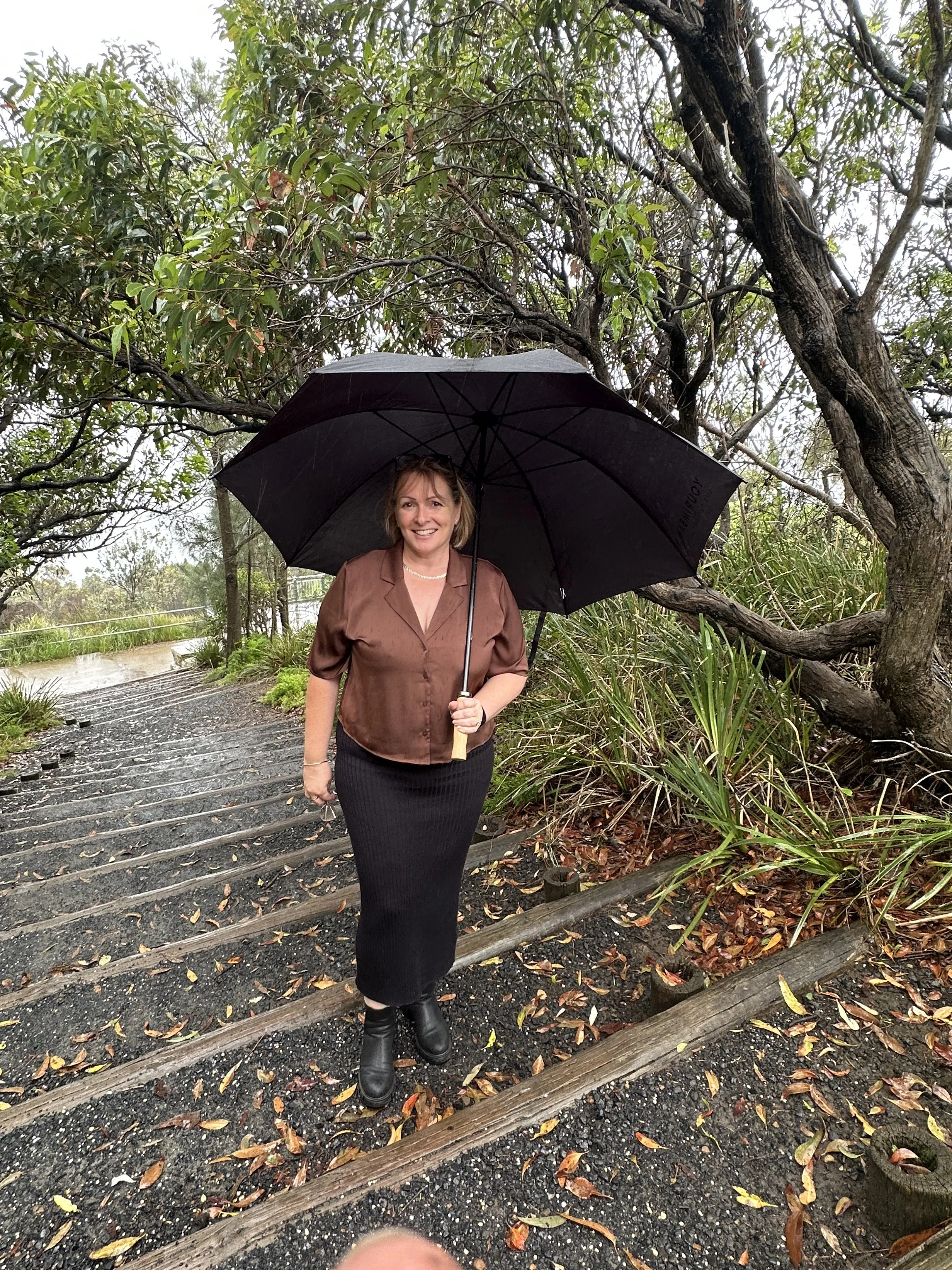Woman standing outdoors on a rainy day holding a black umbrella. She is dressed in a brown blouse and a long black skirt, wearing black boots, and smiling. The background features trees, wet ground, fallen leaves, and wooden logs.