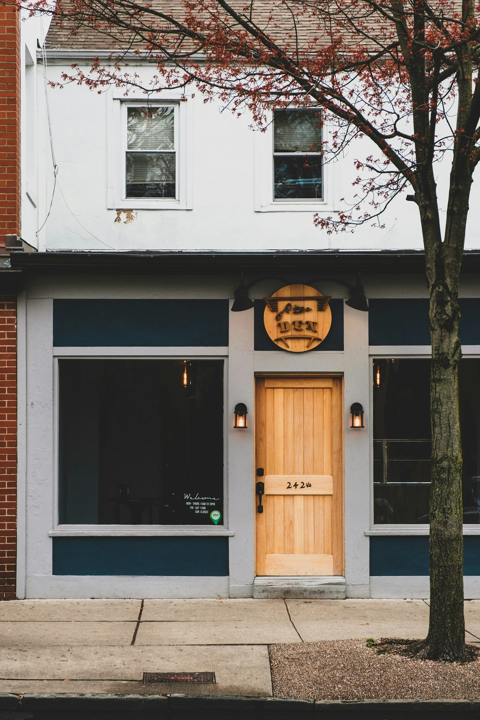 The front view of a modern building with a white exterior, displaying two upper windows, and a ground floor entrance with a wooden door. There is a sign above the entrance that reads 'Jessa DEN' and a smaller wood sign with the number '2426'. Two black light fixtures are mounted on either side of the door. A tree with reddish-brown leaves is on the right side of the image, partially obstructing the view, and the sidewalk and street are visible in the foreground.