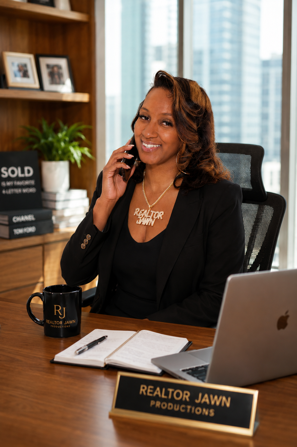 A woman smiling in an office, wearing a black blazer and a necklace that says "Realtor Jawn." She is holding a phone to her ear. On her desk is a black mug with gold lettering that says "Realtor Jawn Productions," a notebook, a pen, and an open laptop. Behind her is a wooden bookshelf with framed photos, books, and a potted plant. Tall office buildings are visible outside the window.