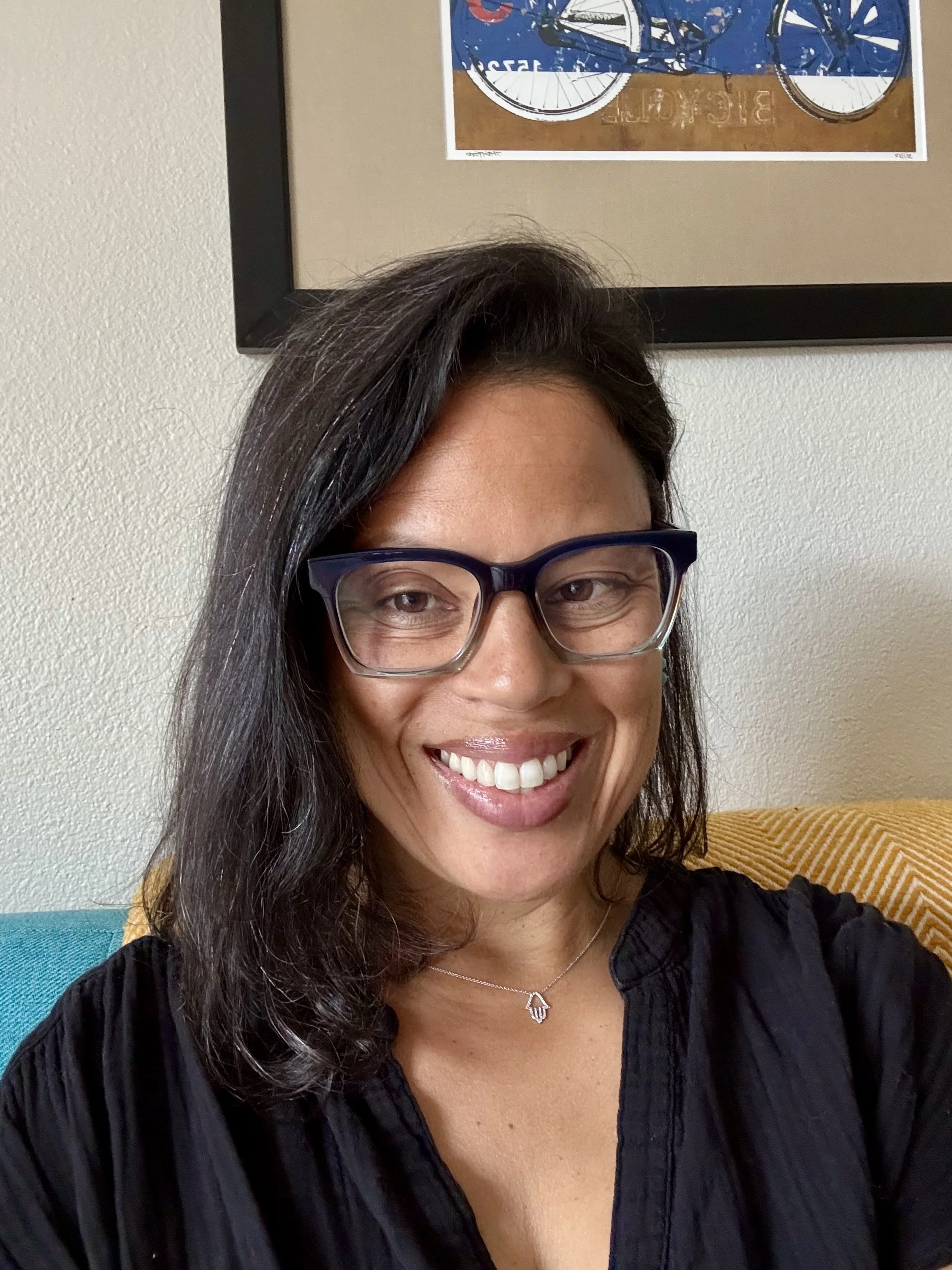 A woman wearing glasses and a necklace smiling for a selfie. She has long dark hair and is dressed in a black top. Behind her are beige walls and some colorful furniture and artwork.