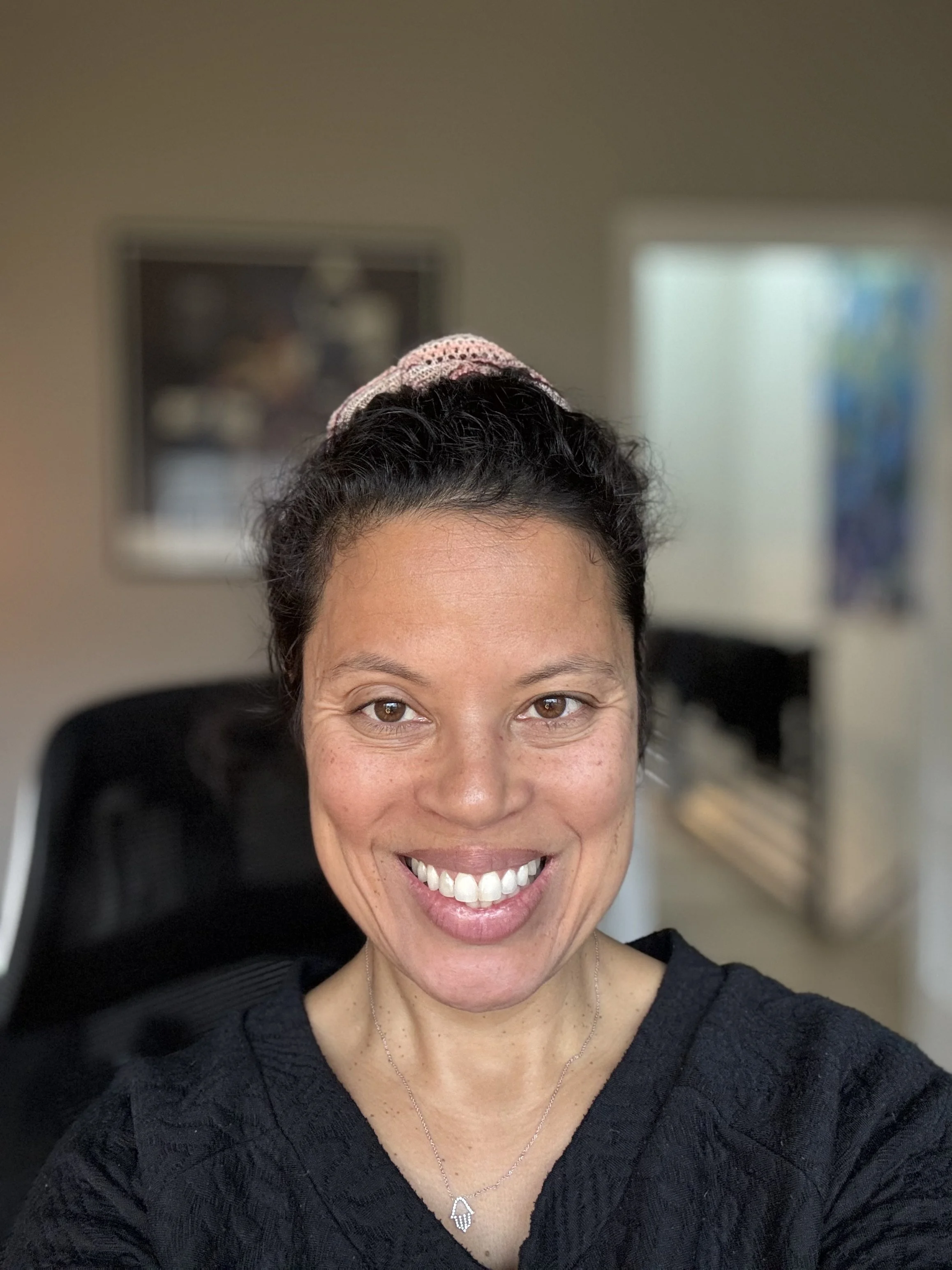 A woman smiling with dark hair, wearing a black top, a silver necklace, and a pink headband with crochet details, in an indoor setting with blurred art on the wall behind her.