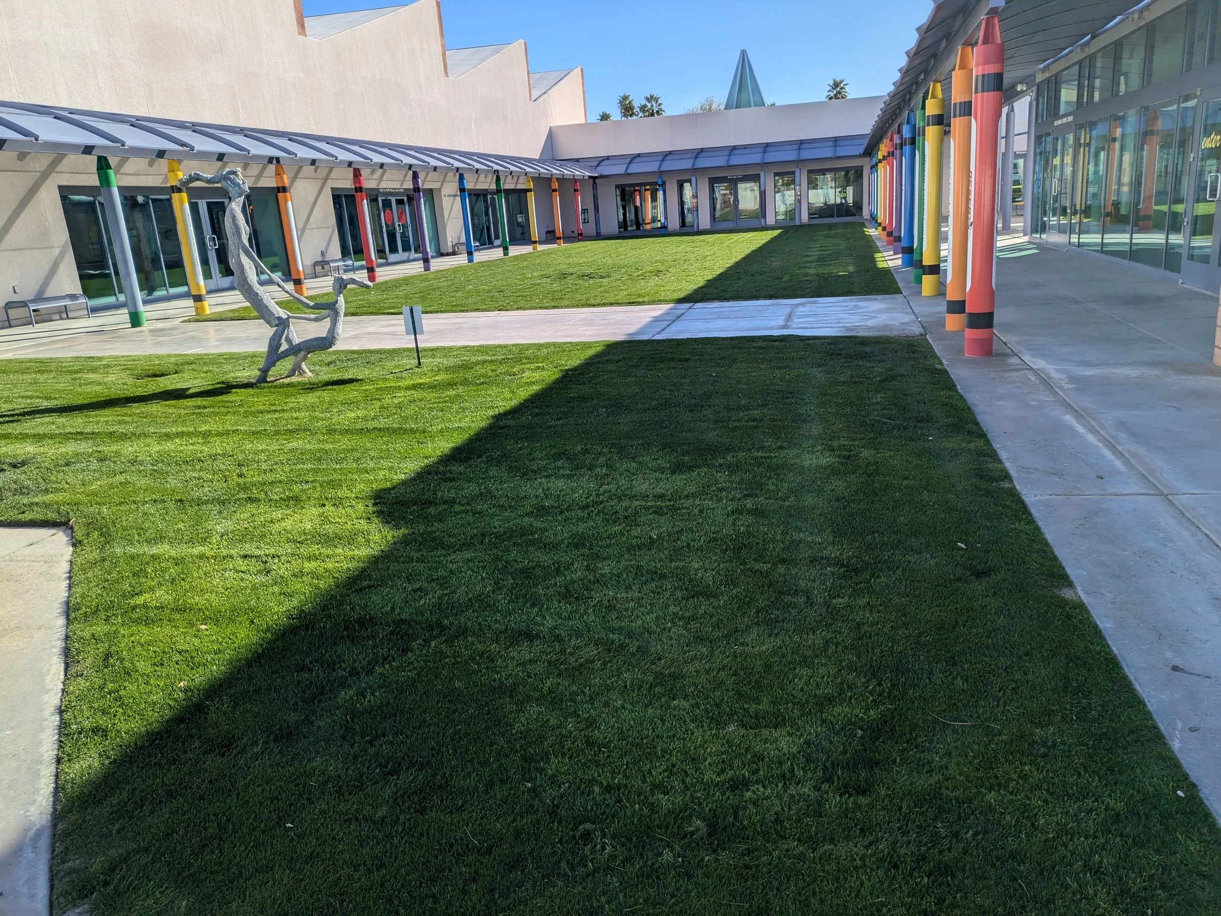 An outdoor area with a green grassy lawn, surrounded by a building with glass walls and colorful vertical poles resembling oversized pencils, and a modern sculpture of a reindeer standing on the grass.
