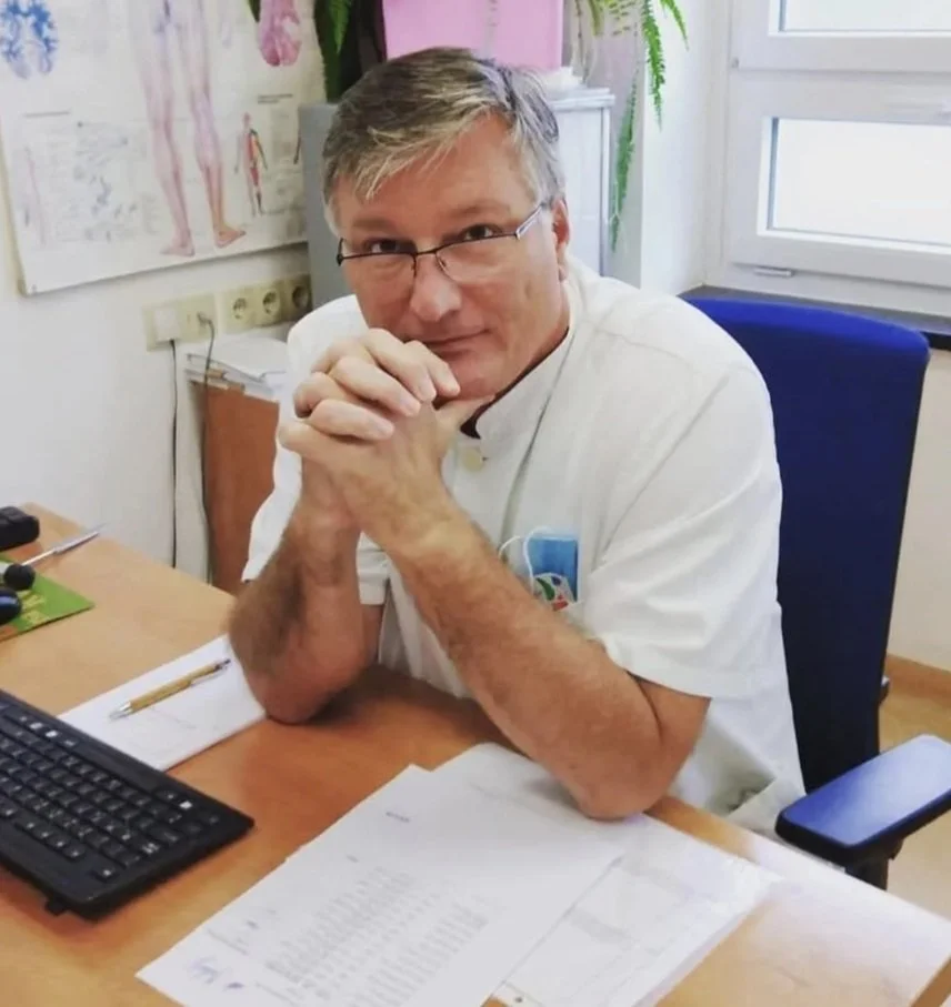 A man in a white medical coat sitting at a desk, hands clasped, looking at the camera, in an office or medical setting with papers, keyboard, and medical chart on the desk.