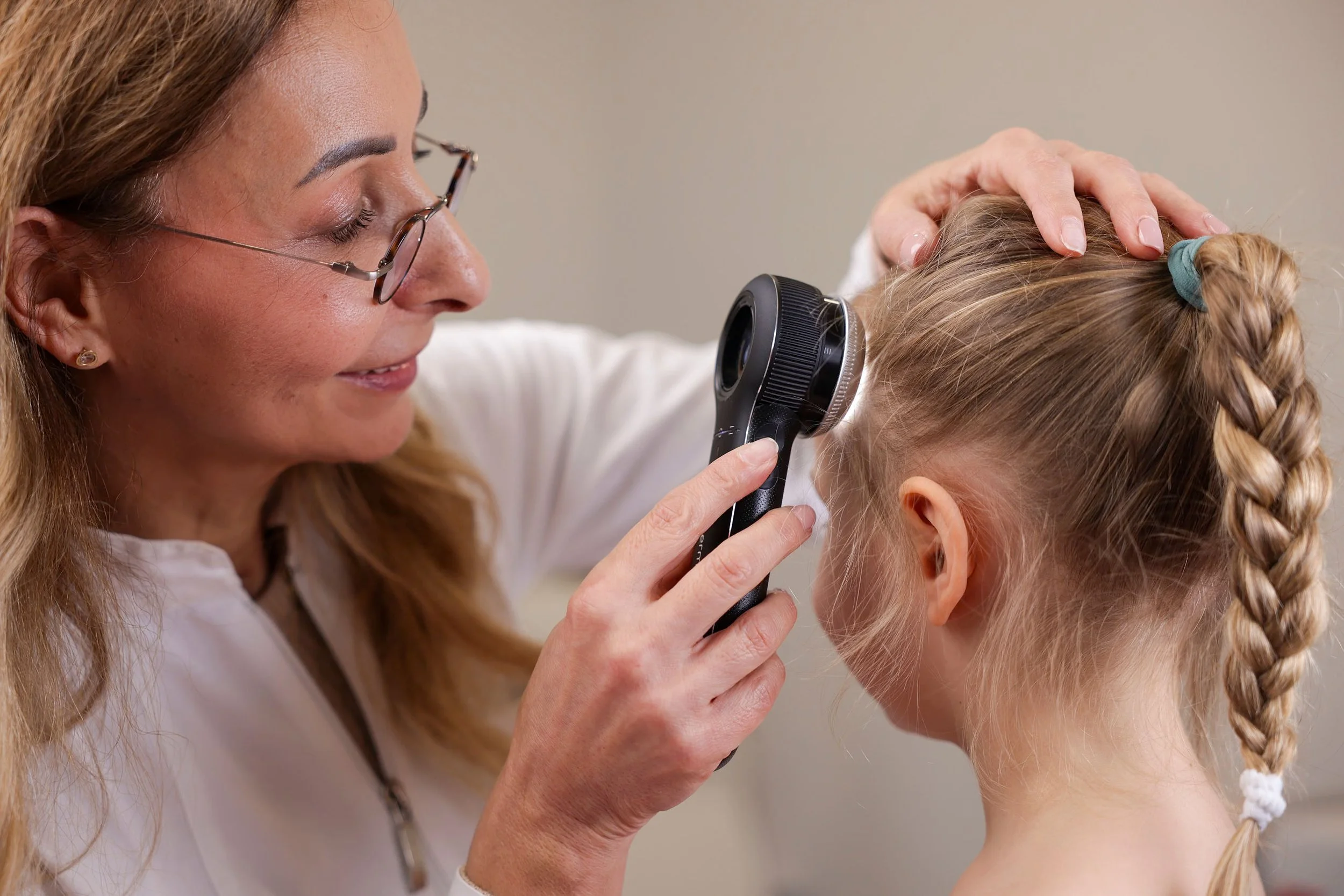 A woman with glasses is examining a young girl's hair with a magnifying glass. The girl has blonde hair styled in a braid.