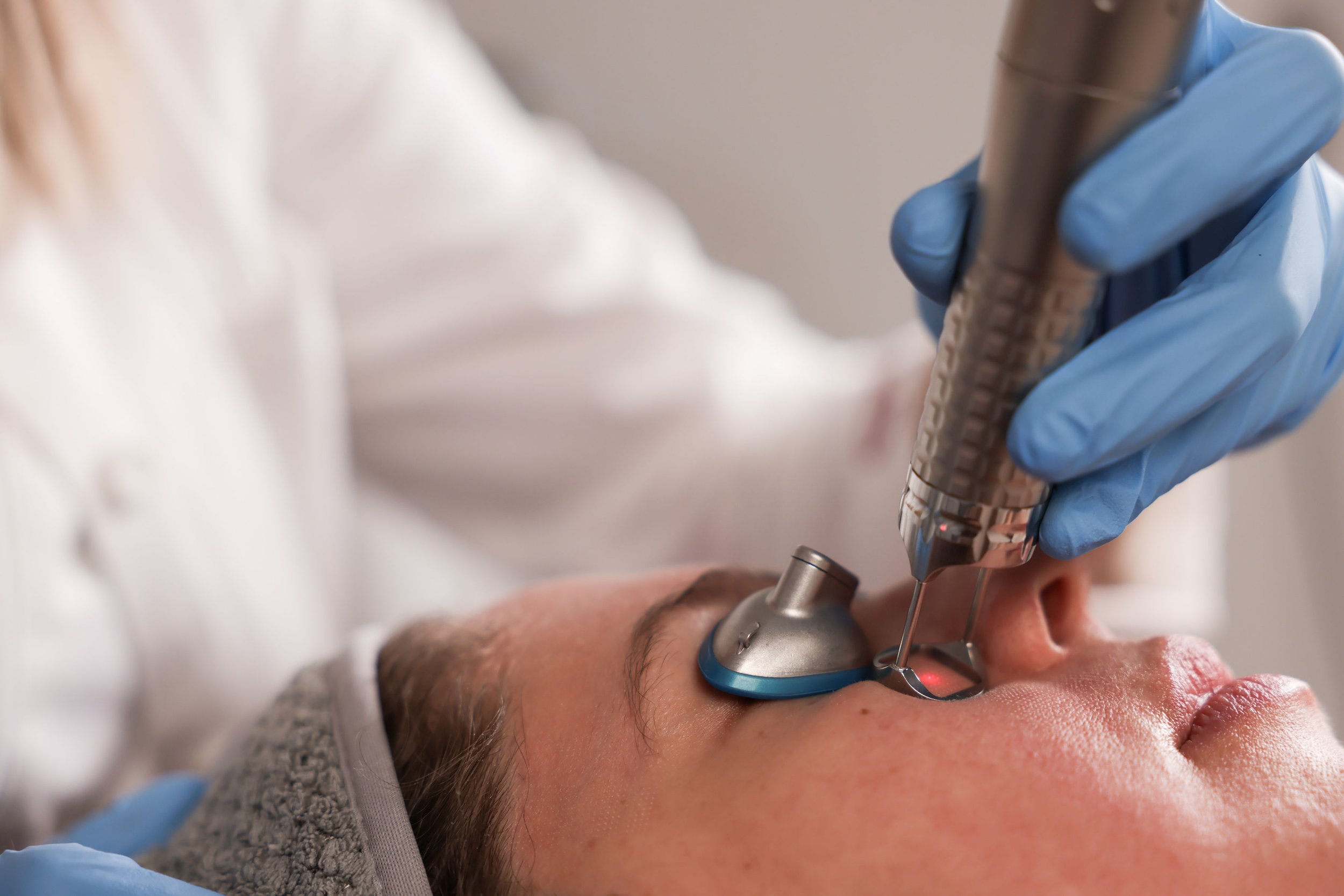 A medical professional wearing blue gloves performs a skin treatment using a handheld device on a man's face, who is laying down with eyes closed.