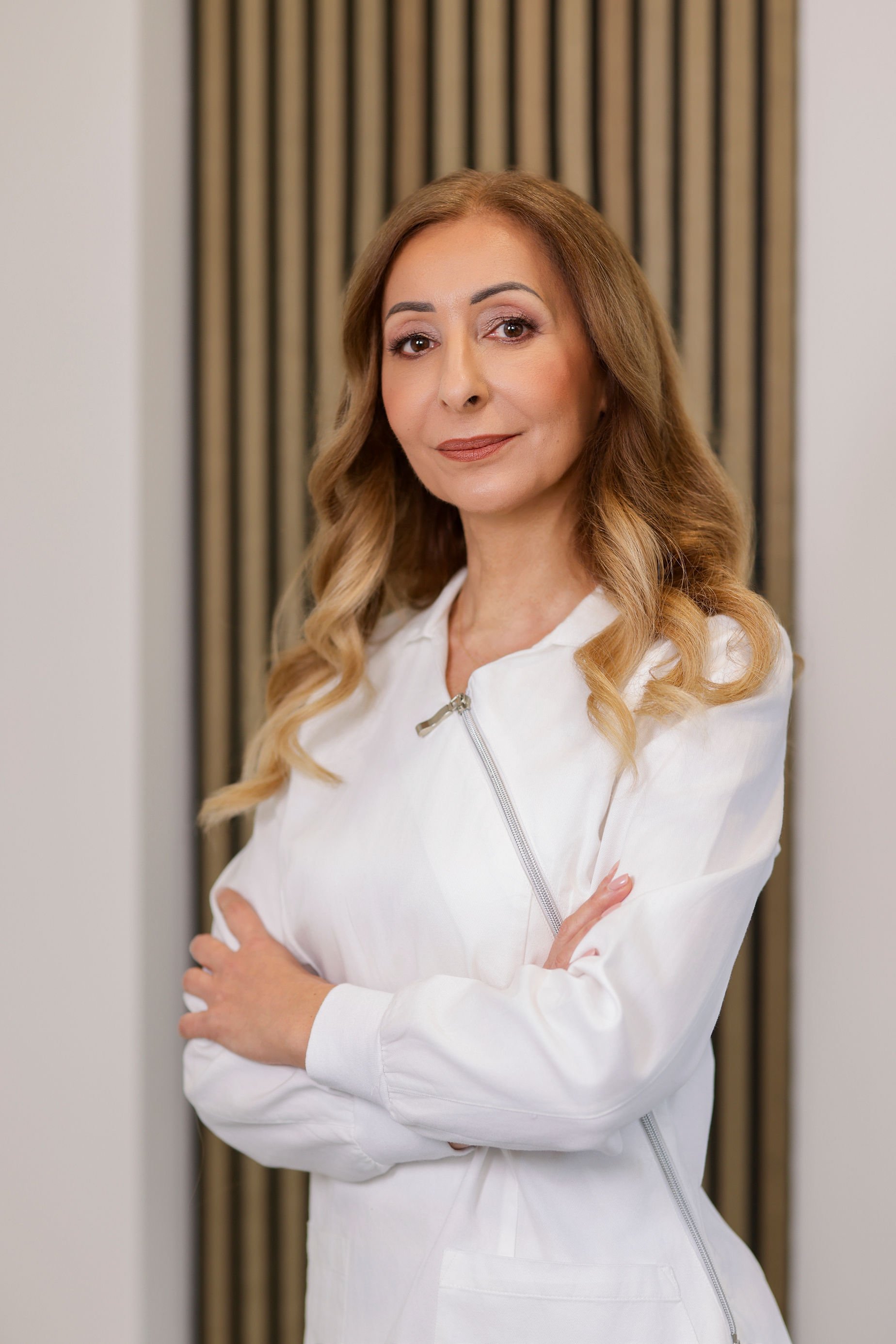 A woman with long wavy blonde hair, wearing a white jacket, standing with arms crossed in front of a striped curtain background.