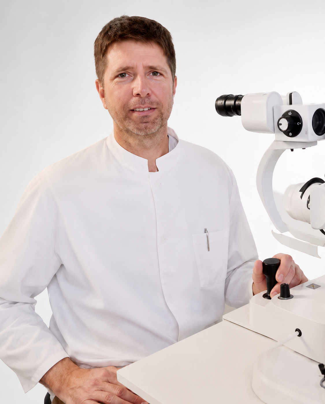 A male scientist in a white lab coat sitting at a table with a microscope.