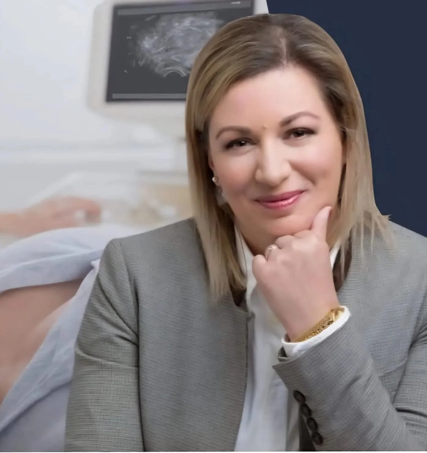 A woman with blonde hair wearing a gray blazer and white shirt, resting her chin on her hand, with a thoughtful expression. In the background, a person in a hospital gown appears to be lying in a hospital bed with medical equipment and an ultrasound image on the wall behind her.
