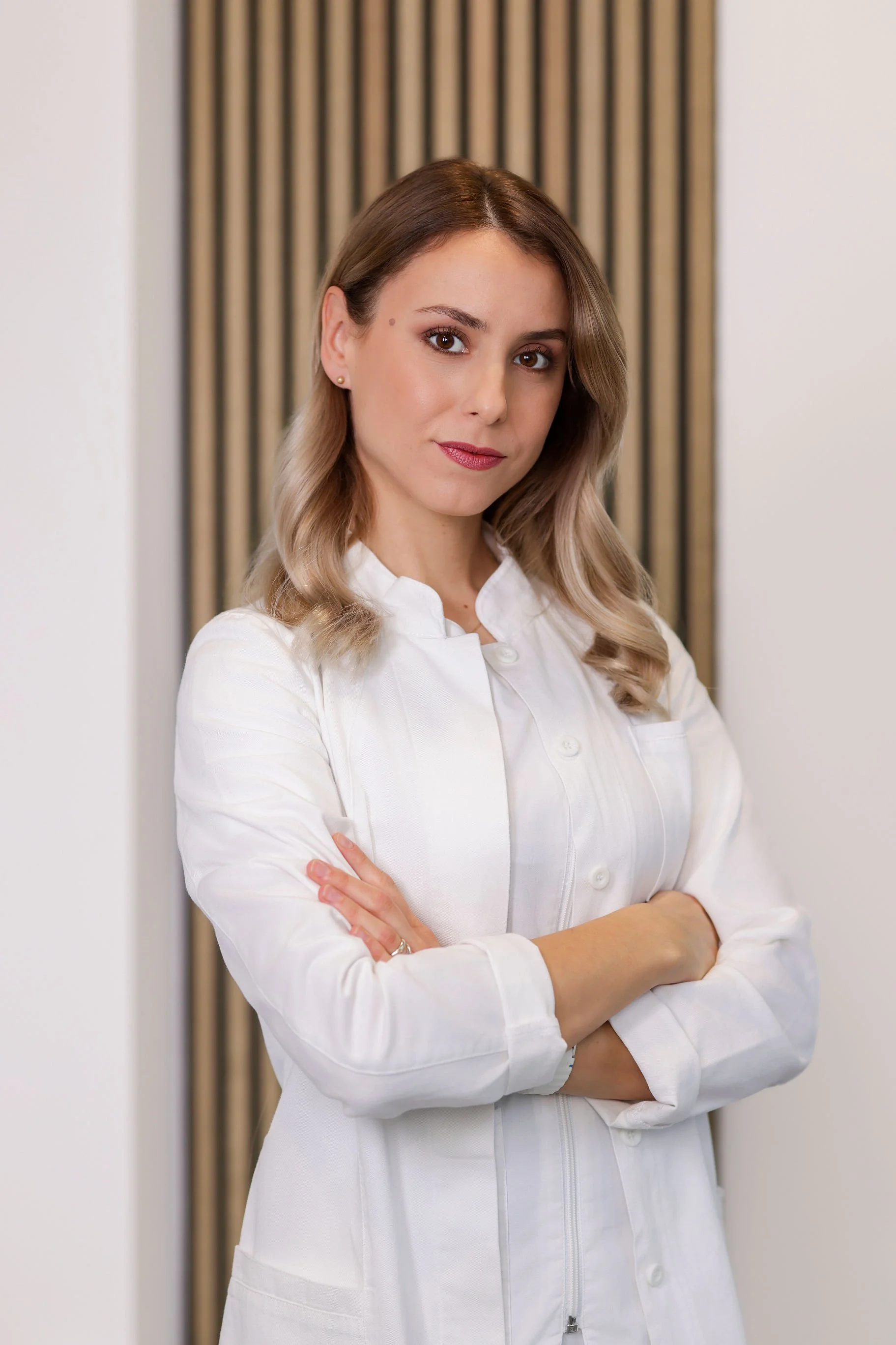 A woman with light brown hair styled in loose waves, wearing a white button-down shirt, standing with arms crossed in front of a neutral wall with vertical striped background.