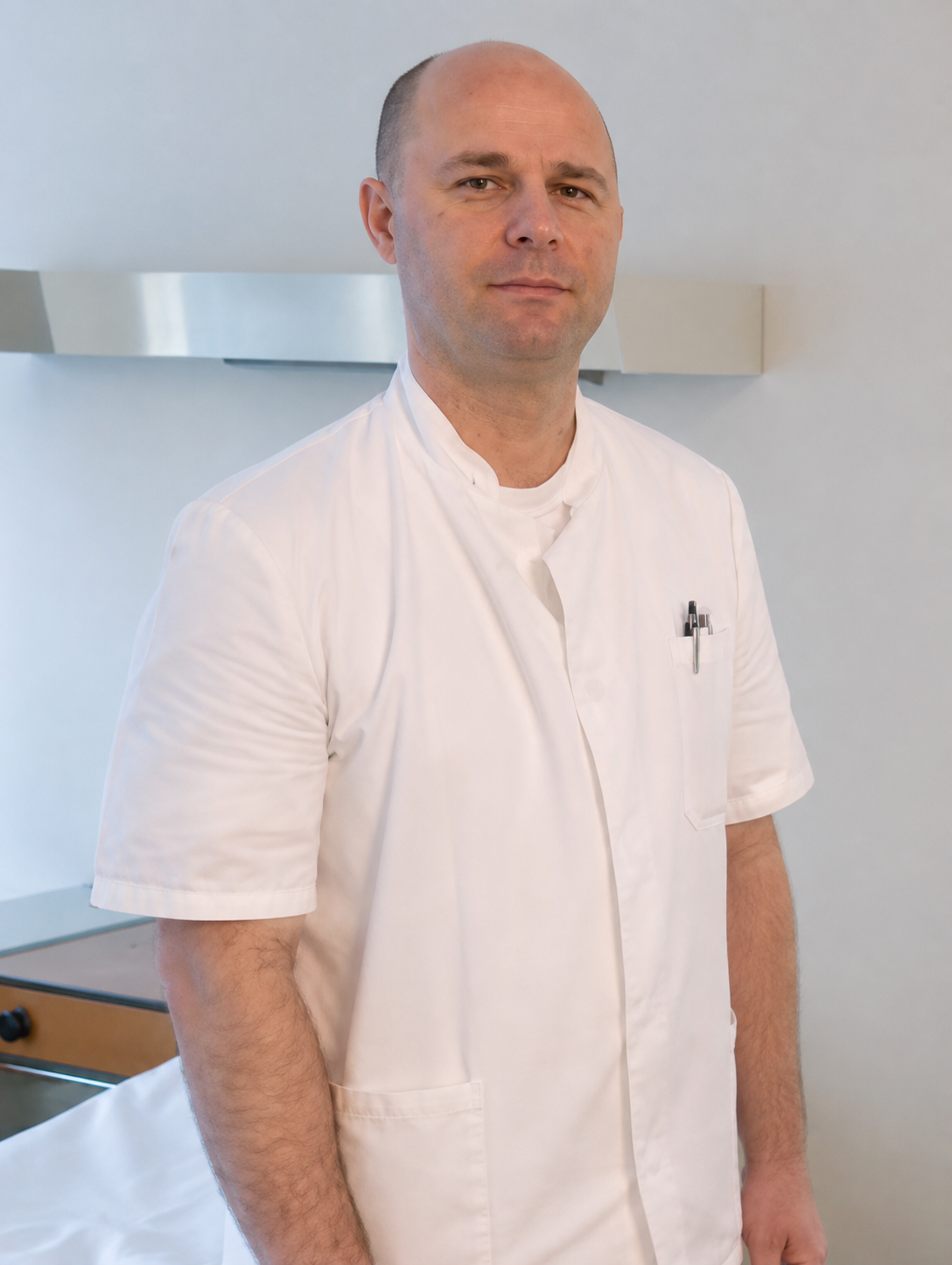 Male healthcare professional wearing white scrubs standing in a clinical setting.