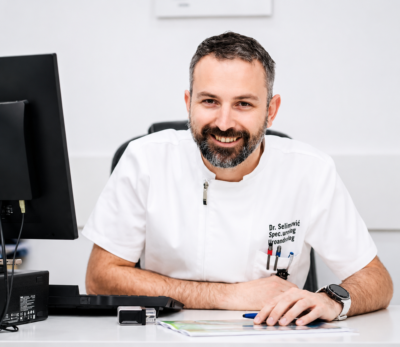 A smiling male doctor with a beard and short hair, wearing a white medical coat, sitting at a desk in an office with a computer monitor, papers, and pens, in a medical or clinical setting.