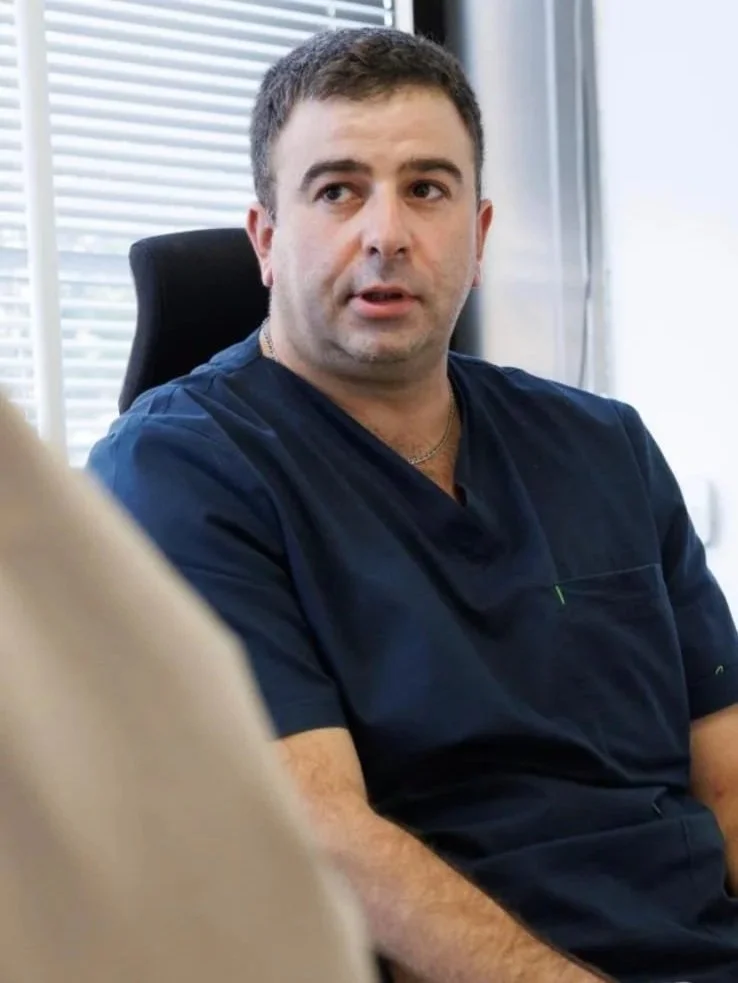 A man with short dark hair and a five o'clock shadow, wearing navy scrubs, sitting in a black office chair, speaking to someone off-camera in a bright office with horizontal blinds.