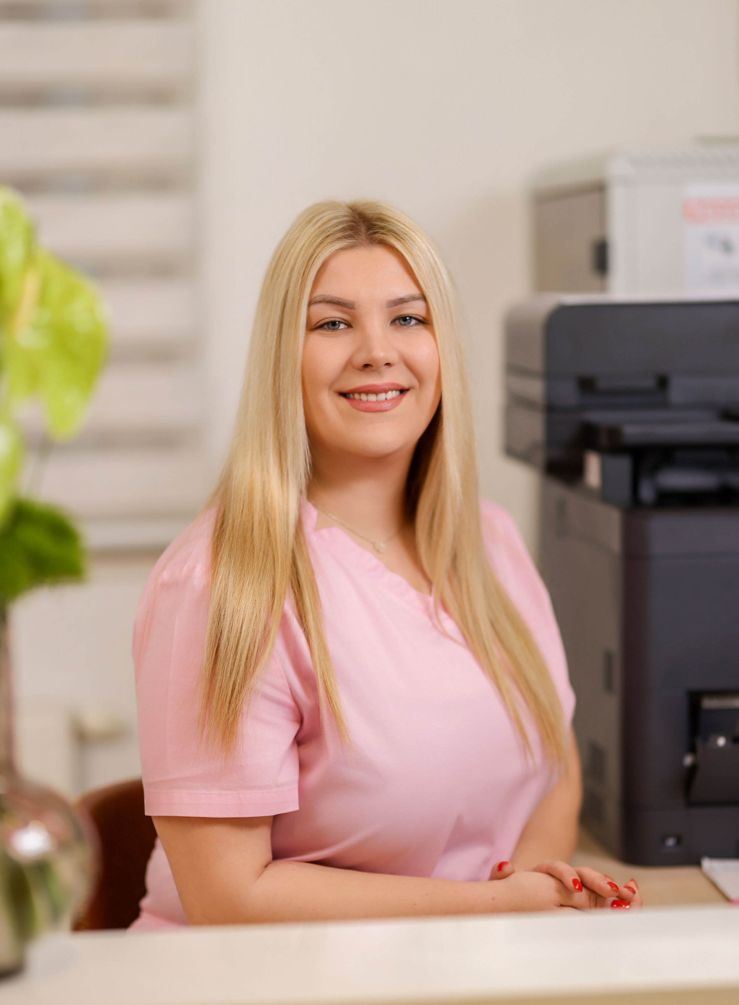 A young woman with long blonde hair, wearing a pink shirt, smiling in an office setting with a printer and white wall in the background.