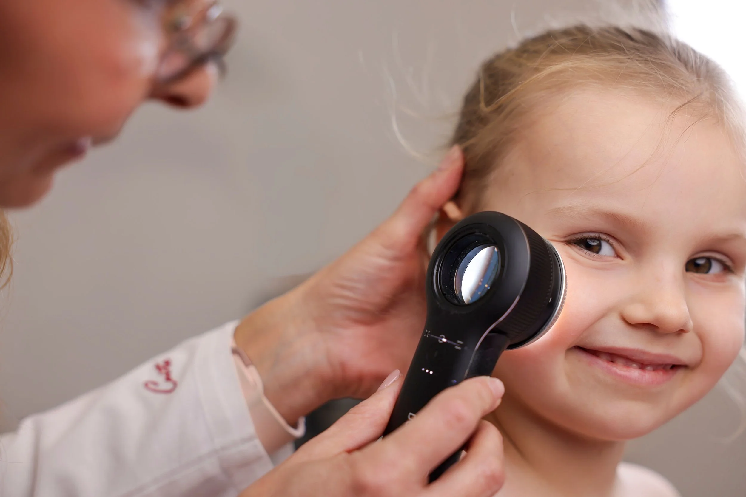 A young girl with brown eyes and light brown hair getting her ears checked with an otoscope by a healthcare provider.