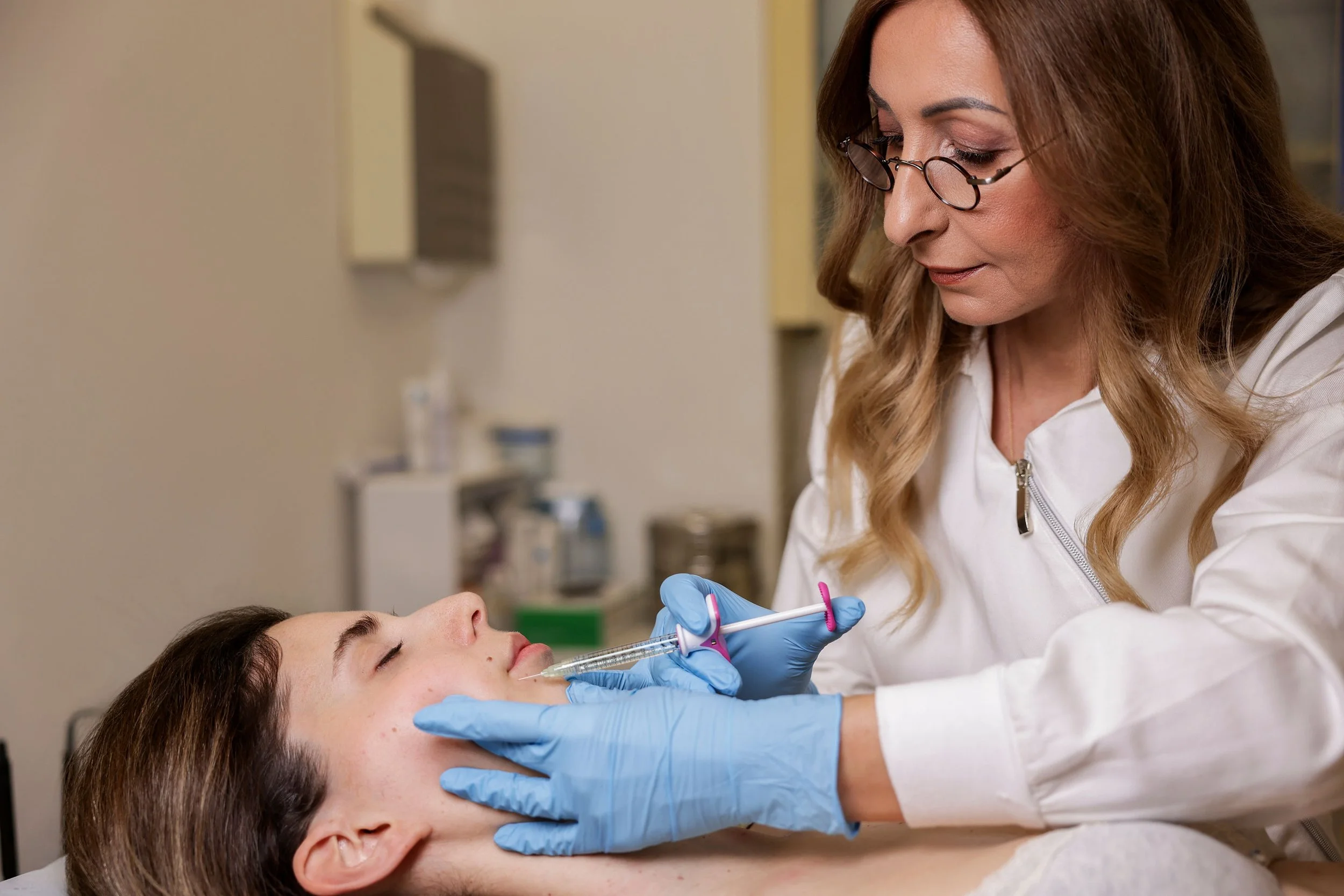 A woman receiving a cosmetic injection from a healthcare professional wearing blue gloves in a medical setting.