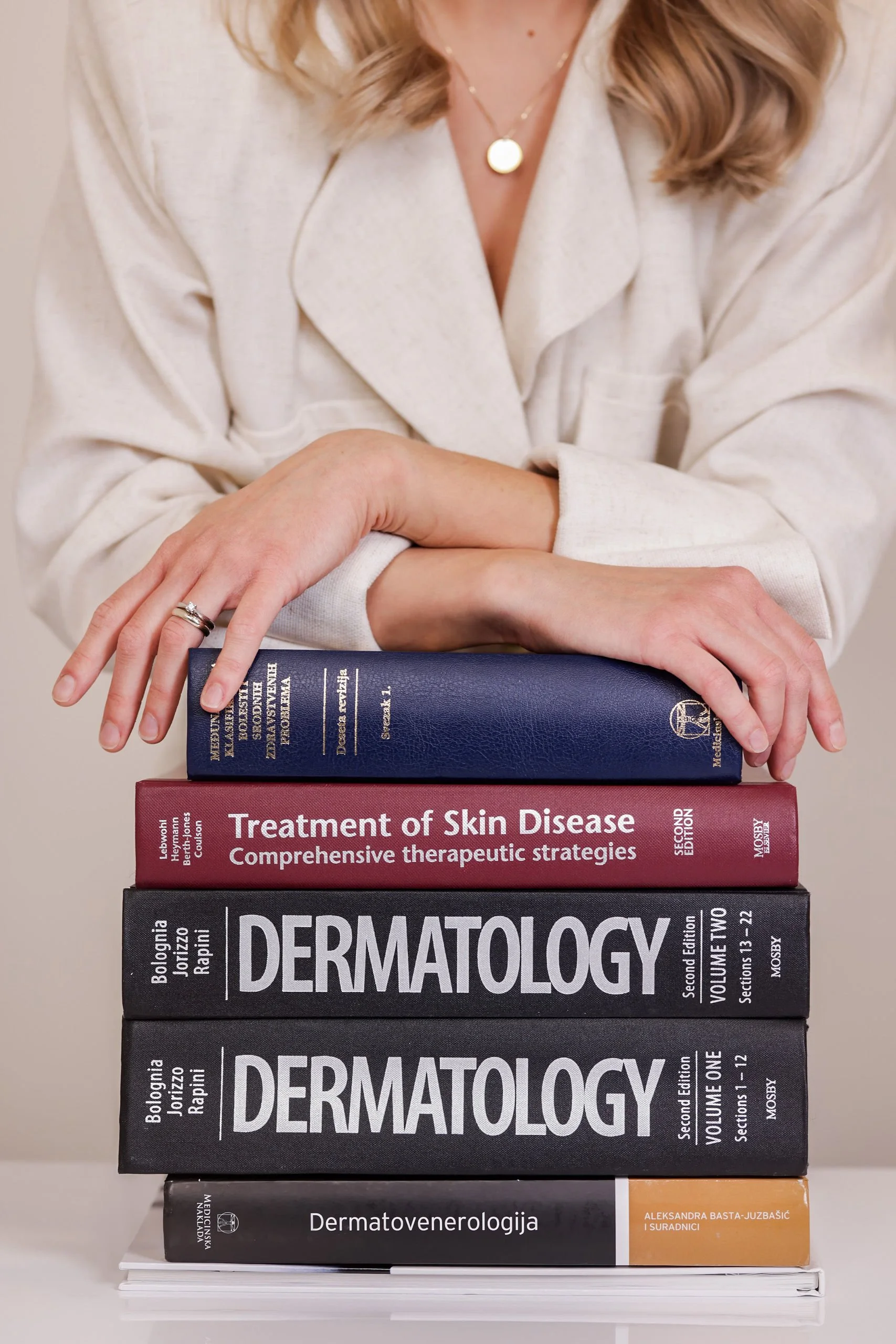 Stack of medical textbooks about dermatology, including titles on skin disease, dermatopathology, and dermato-oncology, topped with a woman in a cream-colored blazer holding the books.