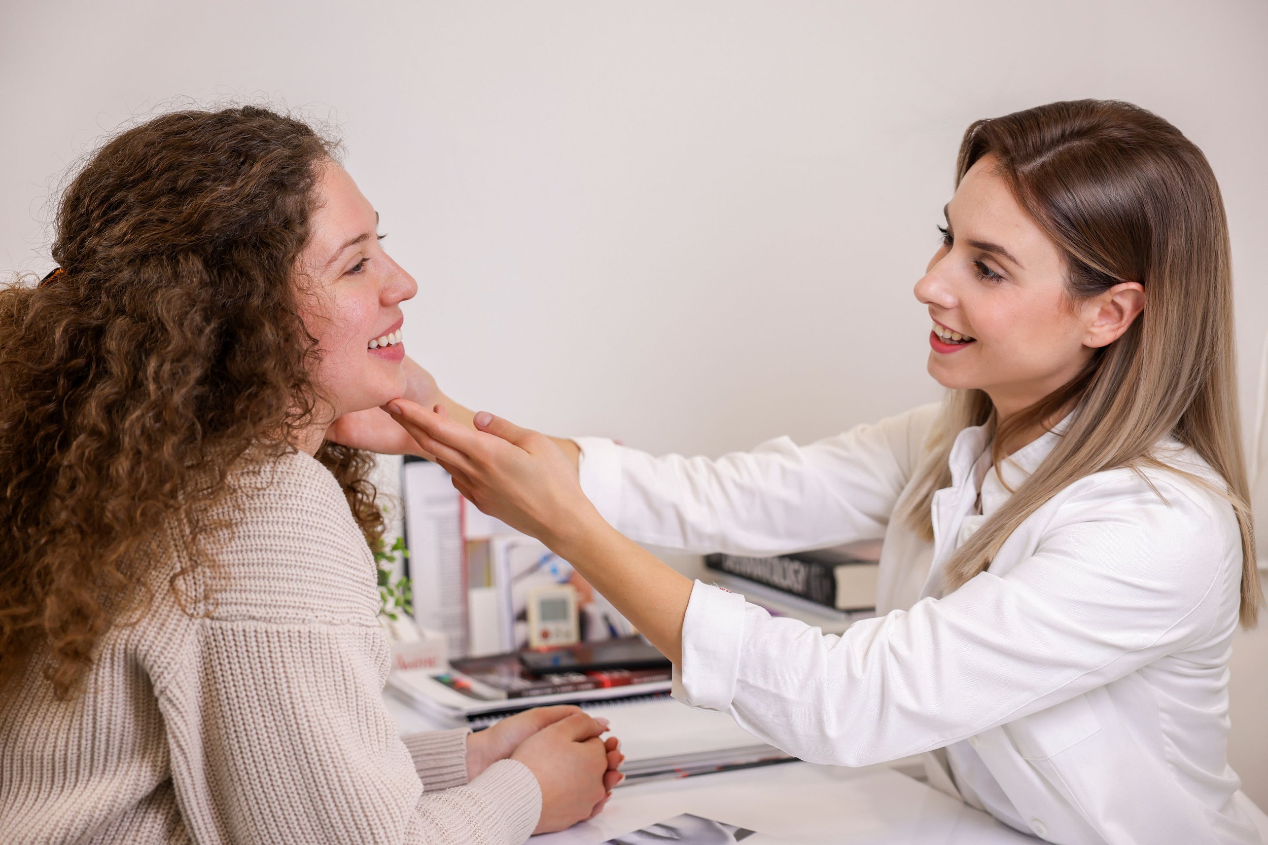 A woman with red hair and a woman with curly brown hair smiling at each other during a consultation in a clinical setting.