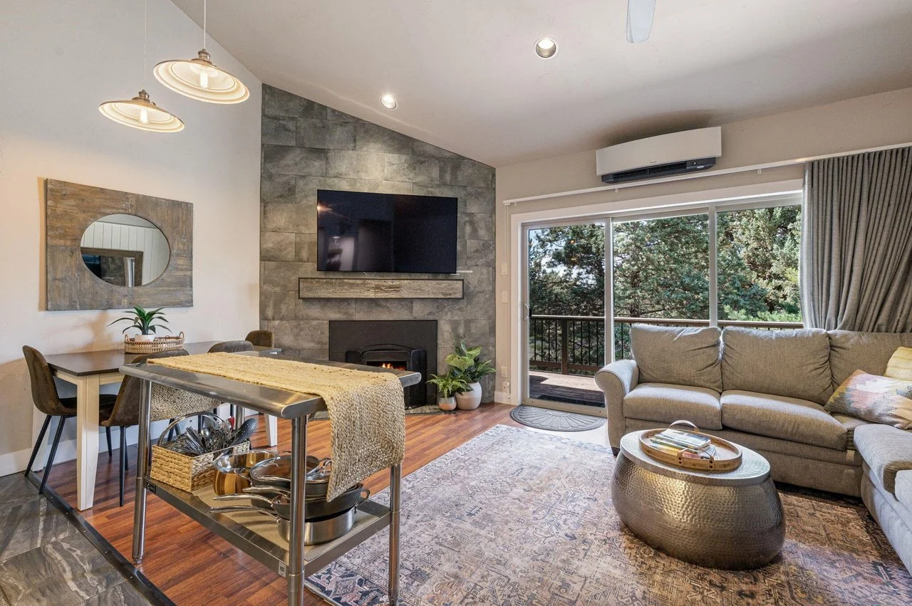 Living room with grey sofa, glass sliding door with curtains, and TV mounted on a stone fireplace wall, with a balcony view of trees outside.