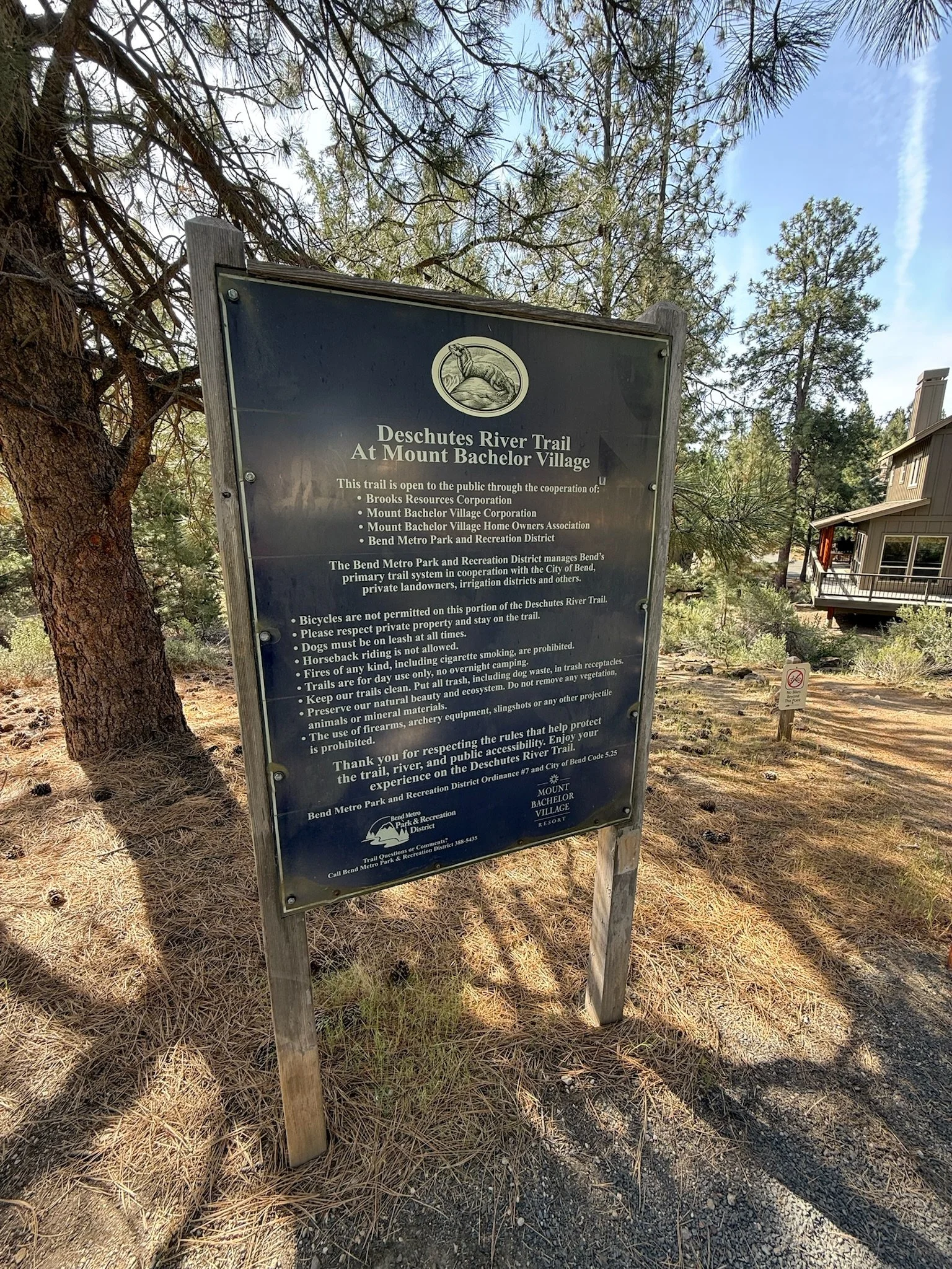 Trail sign for Deschutes River Trail at Mount Bachelor Village, with rules and regulations, surrounded by trees and a house nearby.
