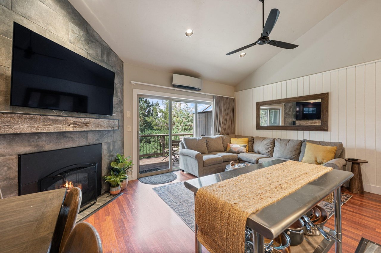 Living room with a fireplace, wall-mounted TV, sliding glass door leading to a balcony, beige sectional sofa, mirror on the wall, ceiling fan, and hardwood floors.