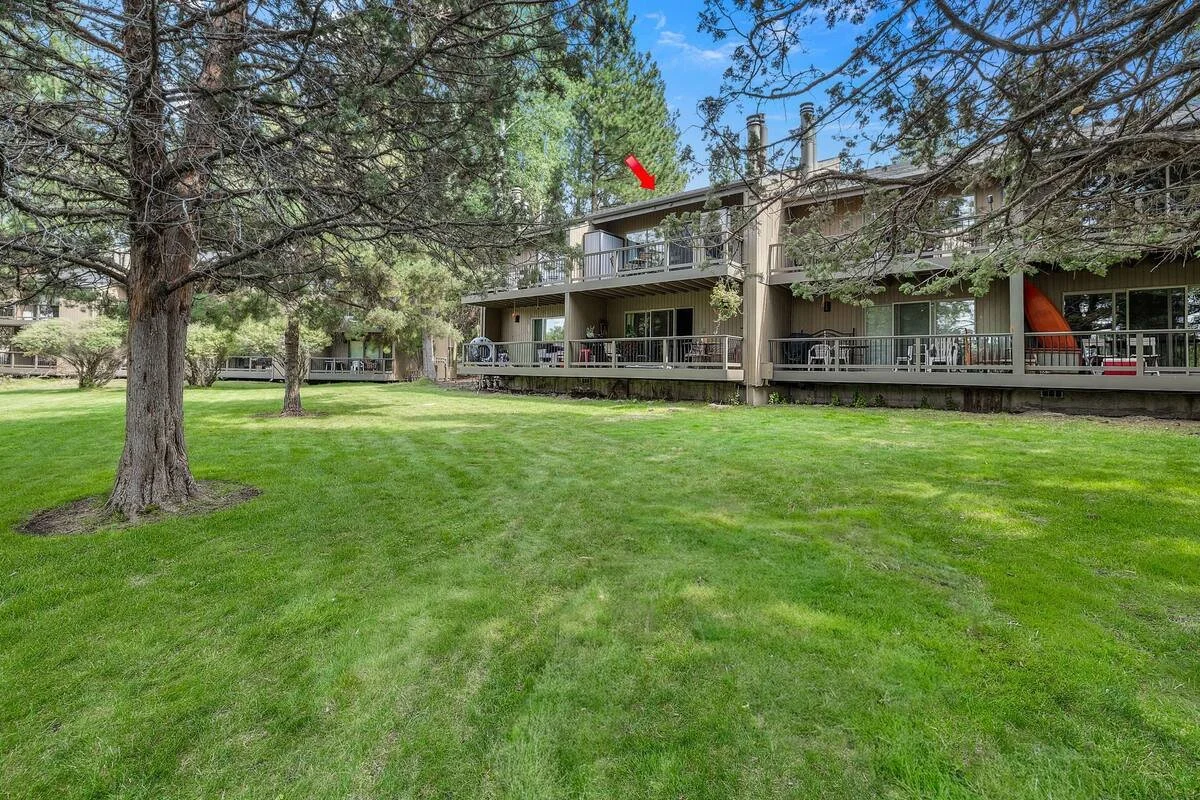A grassy area in front of an apartment complex with trees and a building with balconies, windows, and a slide on the right side.