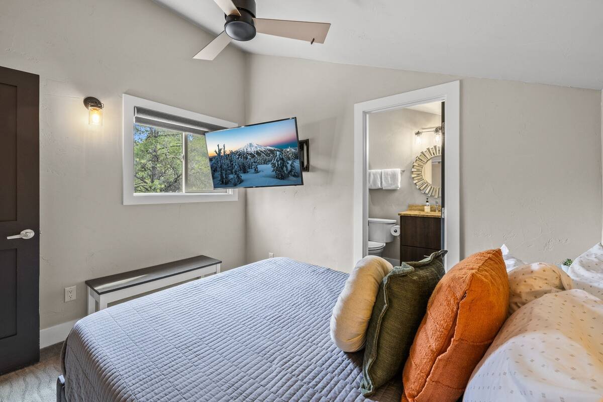 A bedroom with a bed, pillows, a wall-mounted television, a window, and an open bathroom door showing a sink, mirror, and toilet.