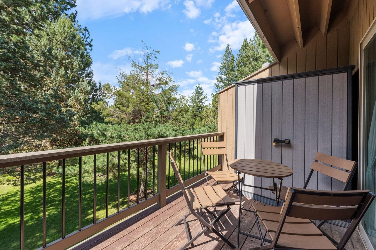 Balcony with four brown metal chairs and a small round table, overlooking a green yard and trees, with a clear blue sky and a few clouds.