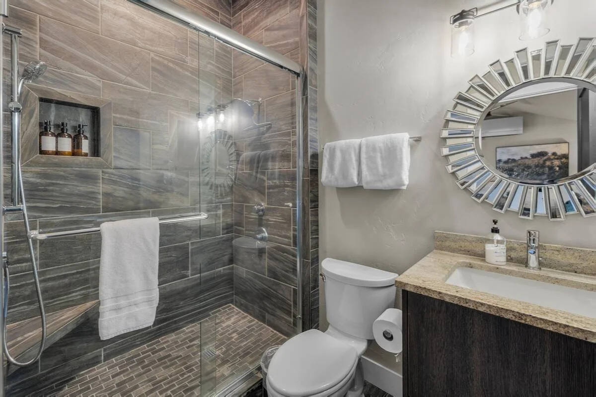 Bathroom with a glass-enclosed shower, a toilet, a granite countertop with a sink, a round decorative mirror, and a towel hanging on the wall.