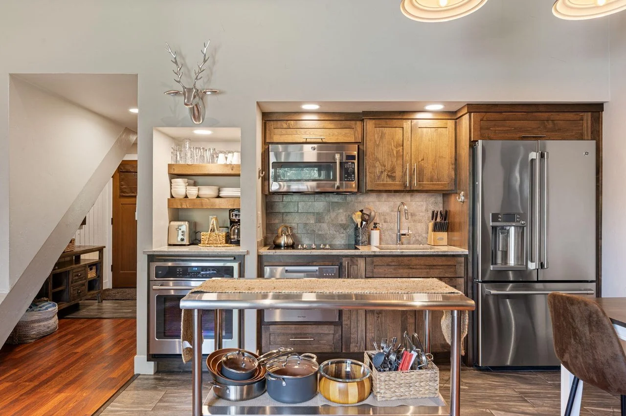 Modern kitchen with wooden cabinets, stainless steel appliances including a microwave and refrigerator, a kitchen island with pots and utensils, open shelves with bowls and glasses, and a decorative deer head wall mount.