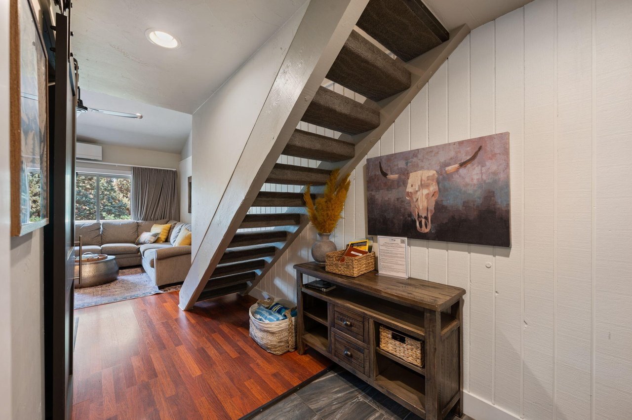Interior view of a living room with a staircase, a beige sectional sofa, a window with curtains, and wooden flooring, with a hallway leading to the staircase area.