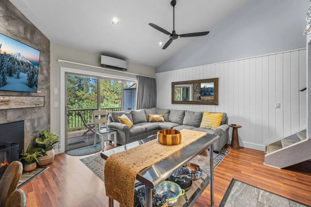 Living room with a gray sofa, sliding glass door leading to a balcony with outdoor seating, wall-mounted TV, wooden and stone fireplace, mirror on the wall, ceiling fan, wood flooring, and various decorative pillows and plants.