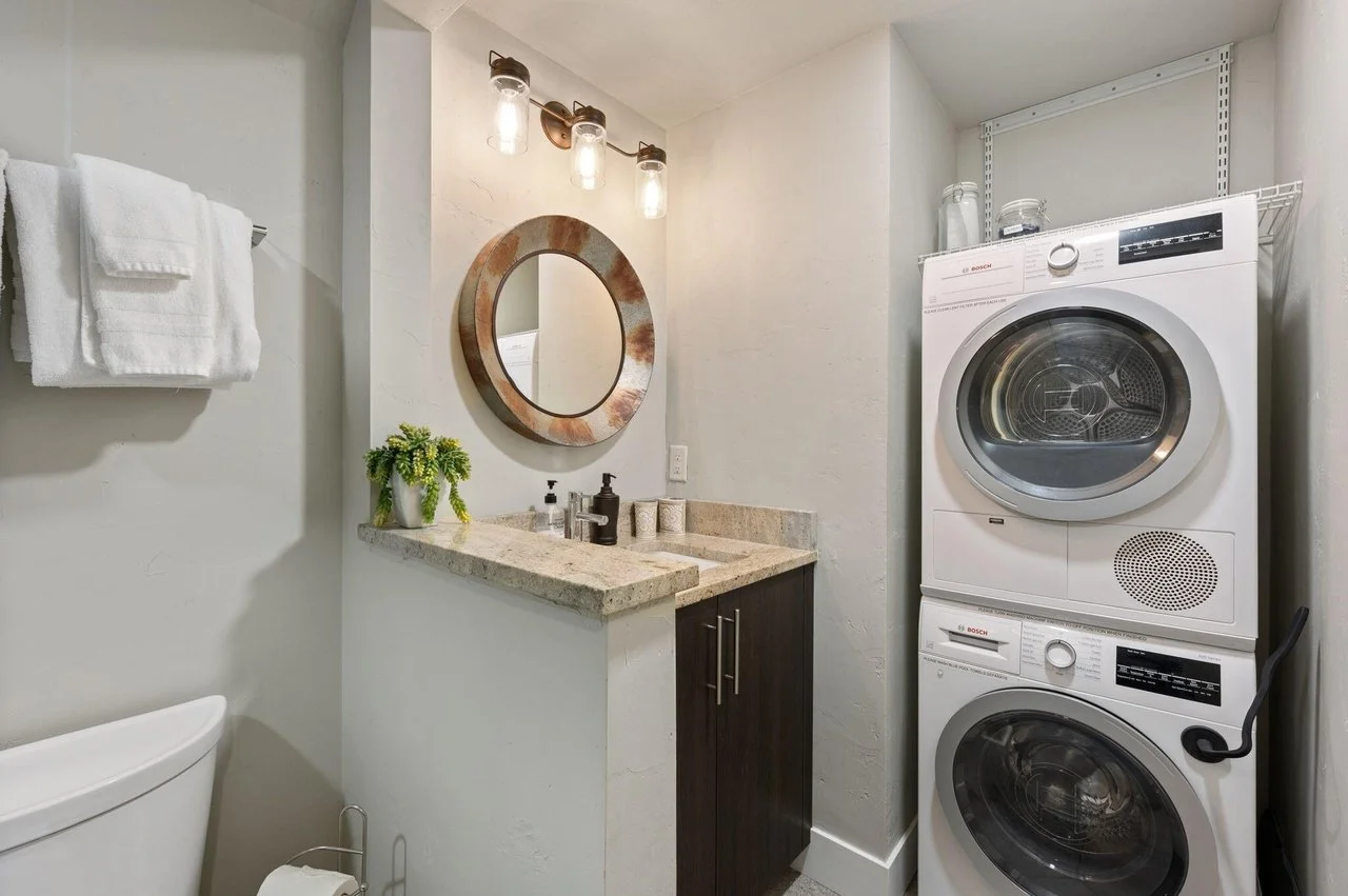 A compact utility bathroom with a stacked washer and dryer, a small vanity with a round mirror, a potted plant, and white towels on a wall-mounted towel rack.