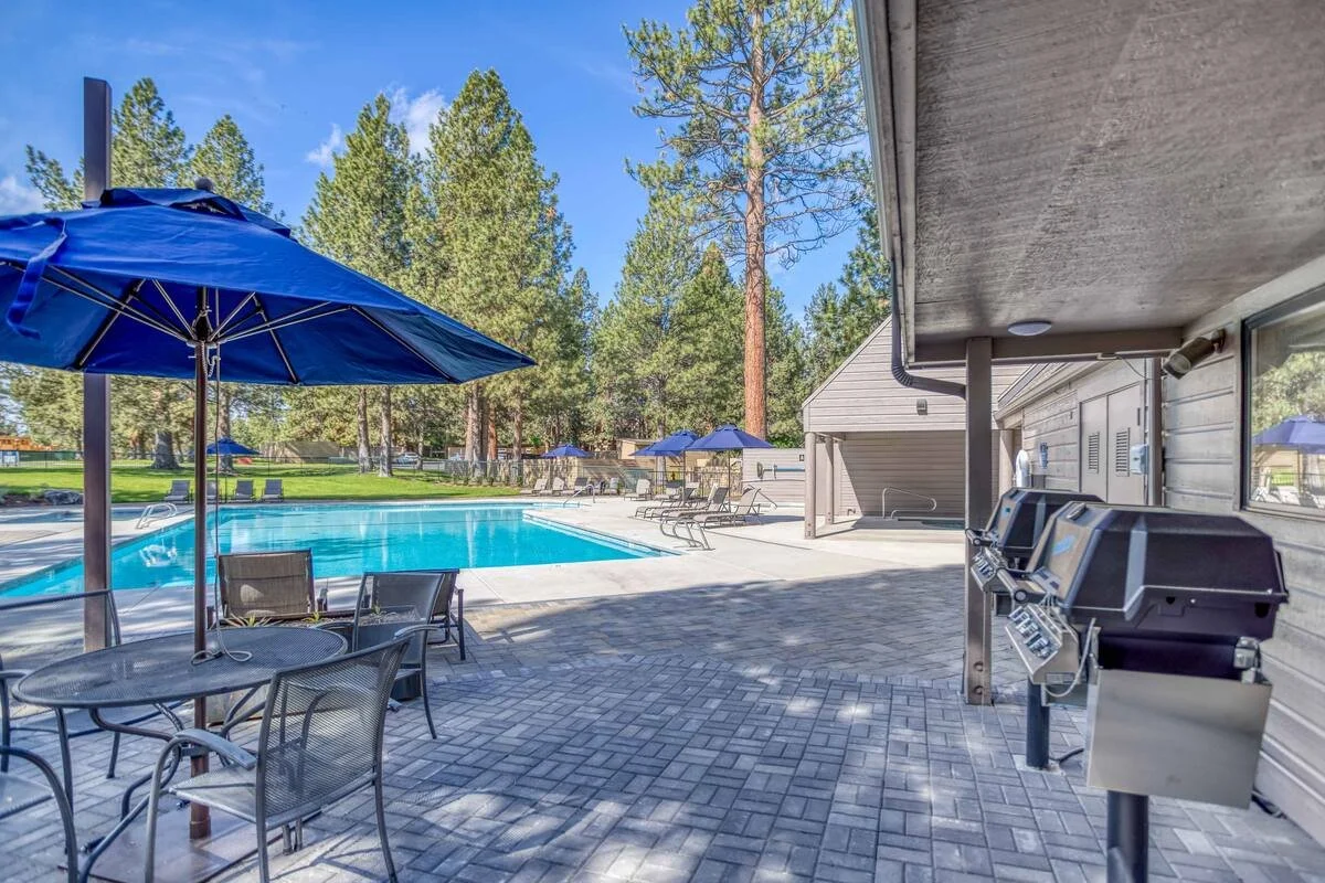 Outdoor swimming pool area with lounge chairs and blue umbrellas, surrounded by trees, with grills and a shaded seating area.
