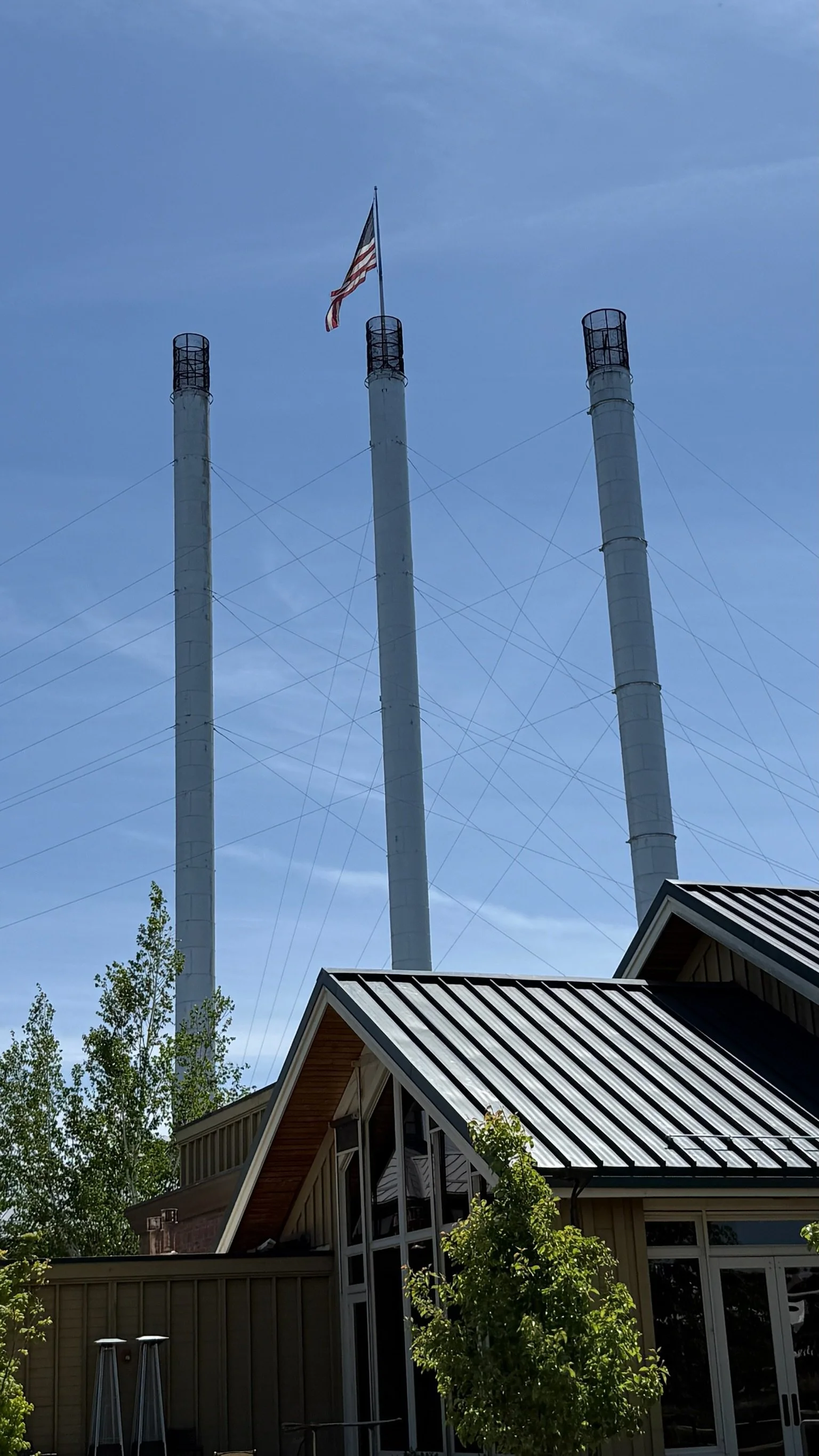 Three large white smokestacks with protective cage tops in the sky, an American flag flying at the top of the middle smokestack, and a house with a metal roof in the foreground surrounded by trees.