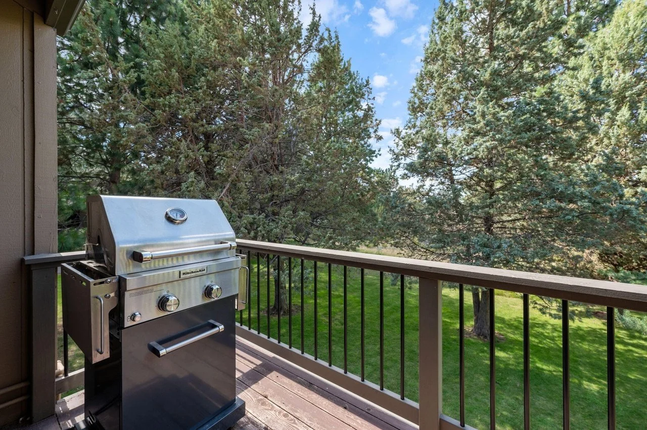 Stainless steel outdoor grill on a balcony overlooking a green yard with large trees and a blue sky.