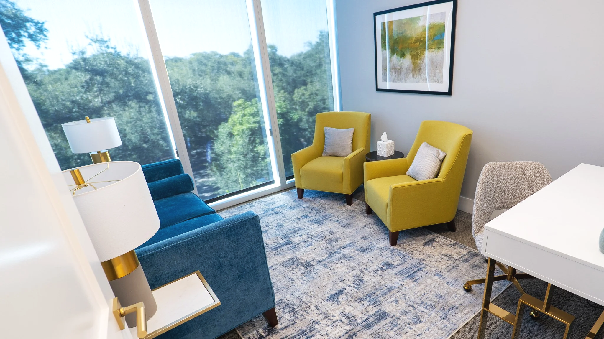 Living room with yellow and blue chairs, large windows showing trees outside, a framed painting on white wall, a white side table with tissue box, and a patterned area rug.