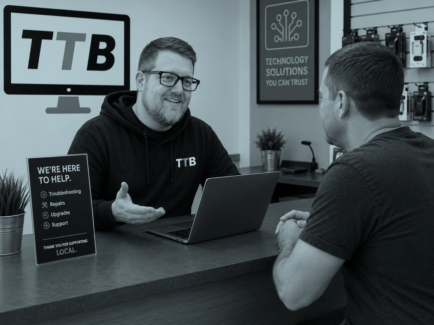 A customer service representative in an office, talking to a customer. The representative is wearing a hoodie with 'TTB' and glasses, smiling and gesturing with one hand. There are signs on the desk offering troubleshooting, repairs, upgrades, and support, and a computer monitor with the 'TTB' logo is visible in the background.