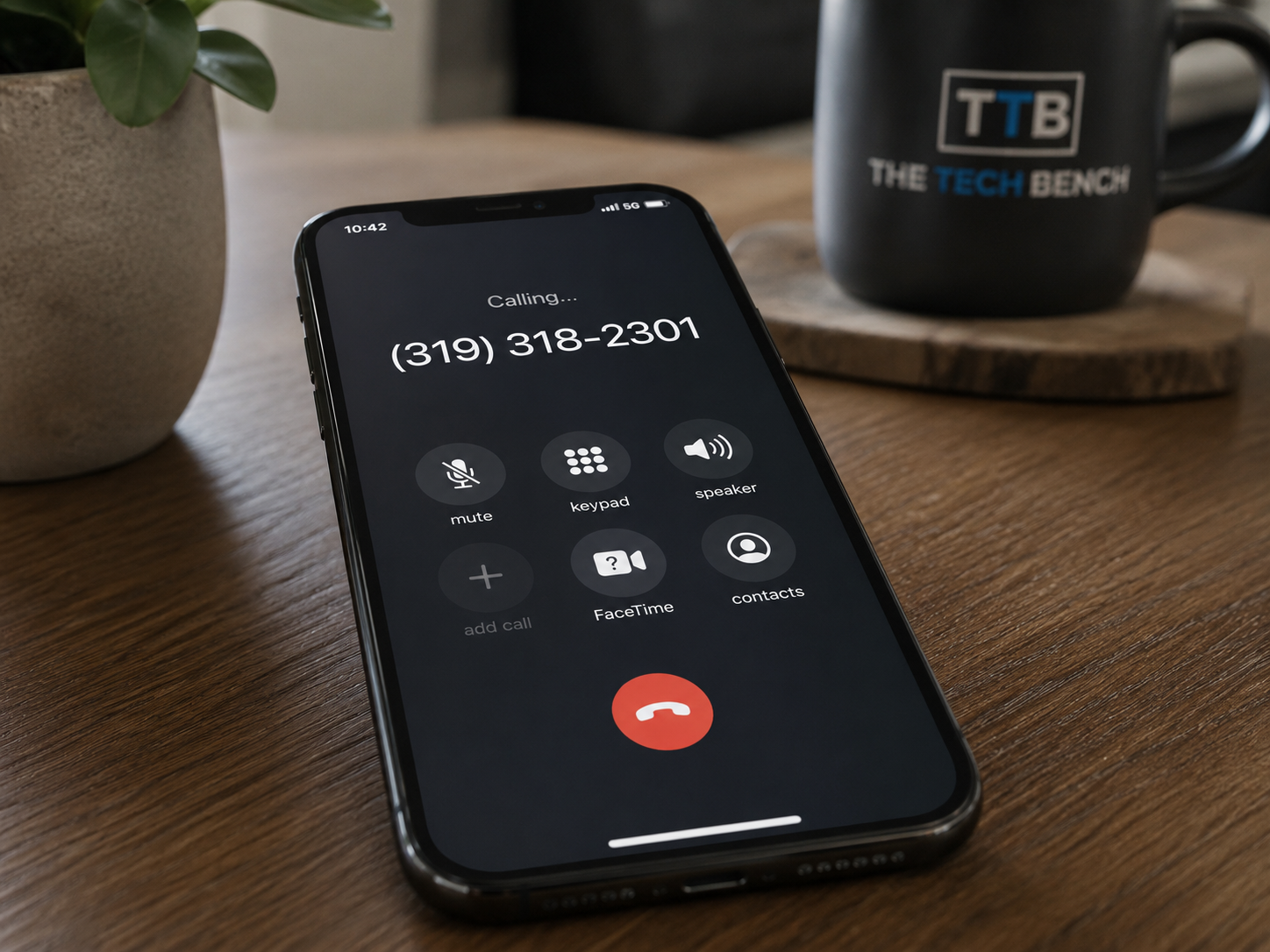 An iPhone on a wooden table during a phone call with the number (319) 318-2301. A potted plant and a black mug with the logo 'TTB The Tech Bench' are also on the table.