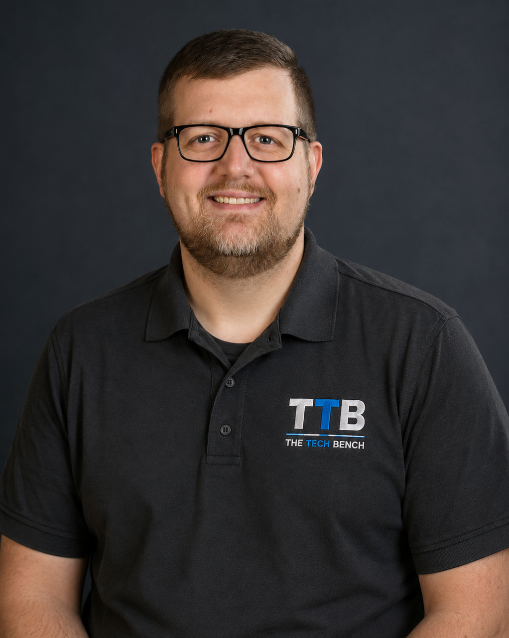 A man with glasses, a beard, and short brown hair smiling in front of a plain dark background, wearing a black polo shirt with a logo that reads "TTB THE TECH BENCH".