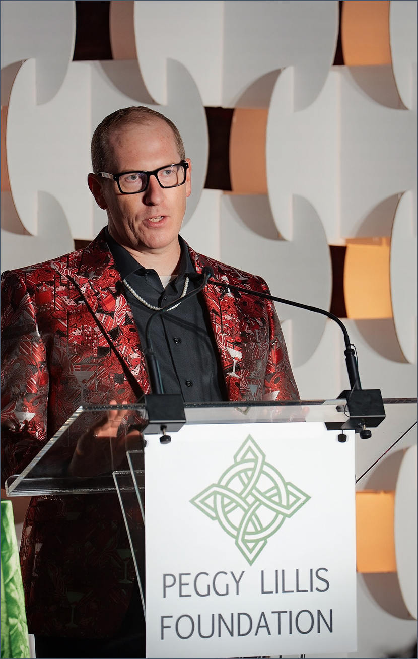 A man wearing glasses and a red patterned blazer speaking at a podium with a Peggy Lillis Foundation sign.