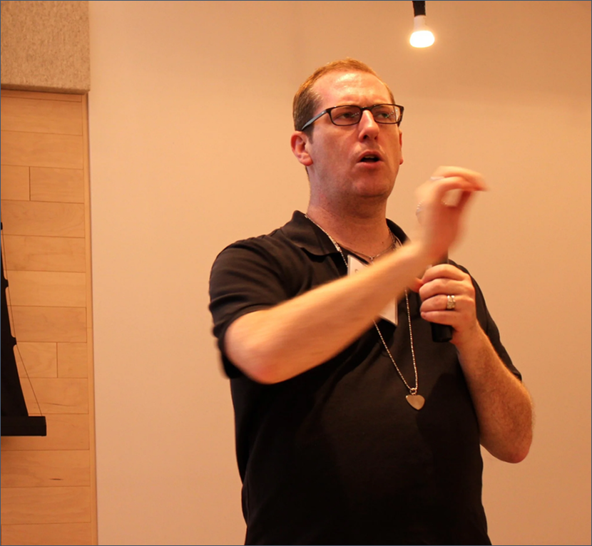 A man with glasses, wearing a black polo shirt, holding a wireless microphone, speaking, with a necklace that has a heart-shaped pendant, gesturing with his right hand in a room with wooden paneling on the wall and a ceiling light.